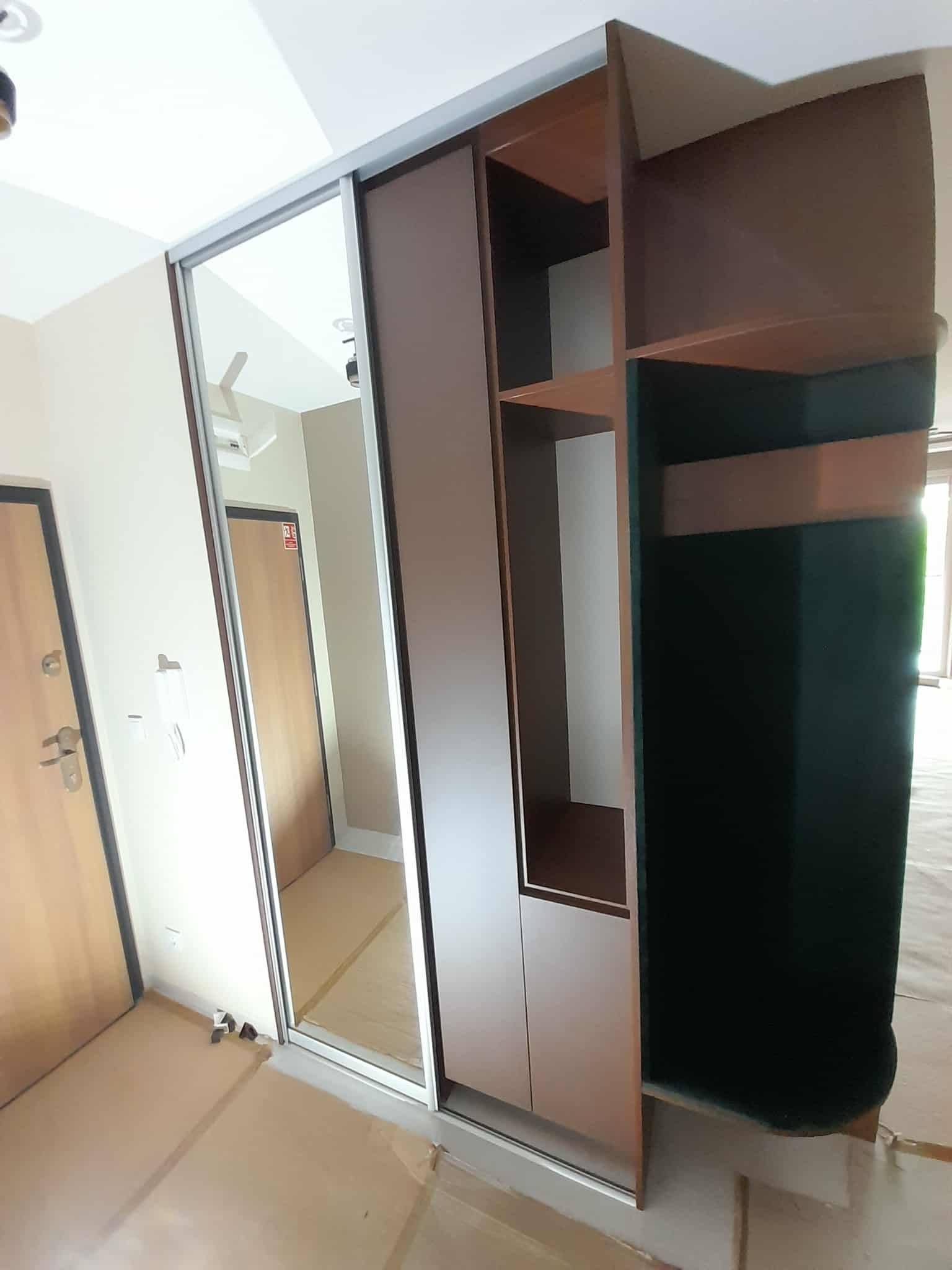 Szafa wnękowa z lustrzanymi drzwiami przesuwnymi i otwartą częścią z półkami, widoczna w trakcie montażu, zabezpieczona kartonem na podłodze.