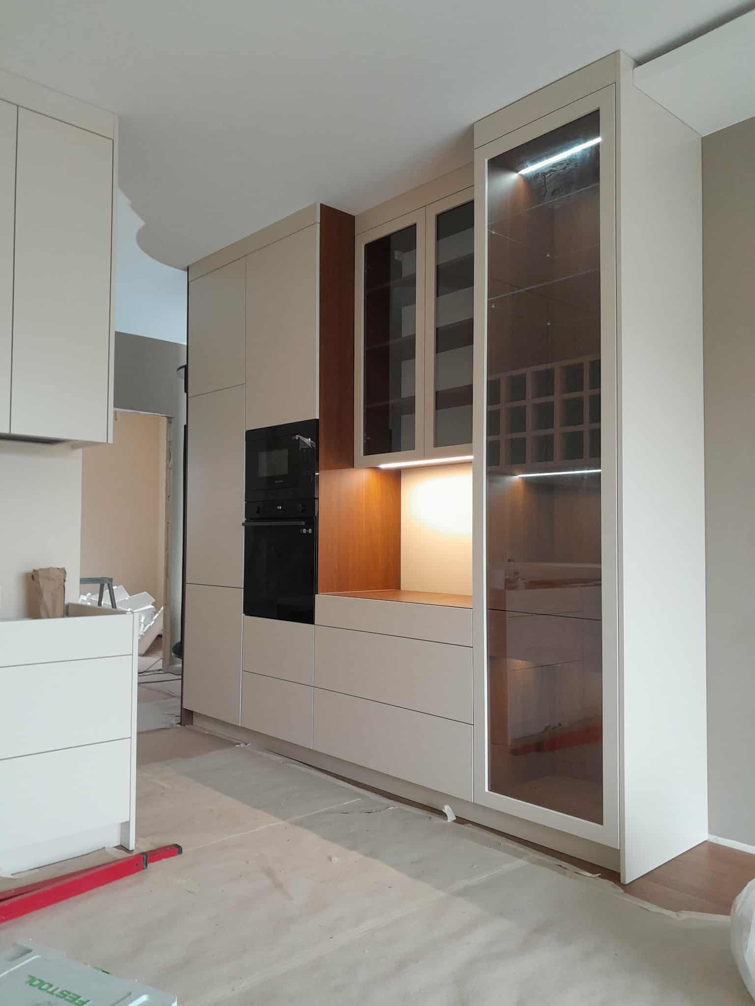 Jasna kuchnia z zabudową, czarnym piekarnikiem i witryną z podświetleniem LED. Widoczne narzędzia na podłodze sugerują montaż.