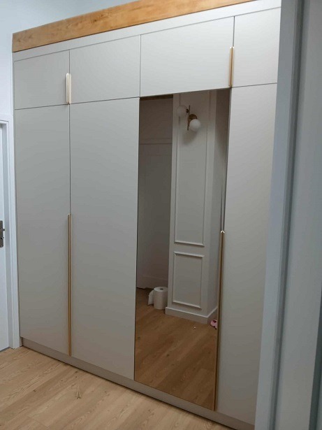 Jasna szafa wnękowa z lustrem, złotymi uchwytami i drewnianą nadstawką, wkomponowana w jasne wnętrze. Widoczne detale wykończenia ścian.