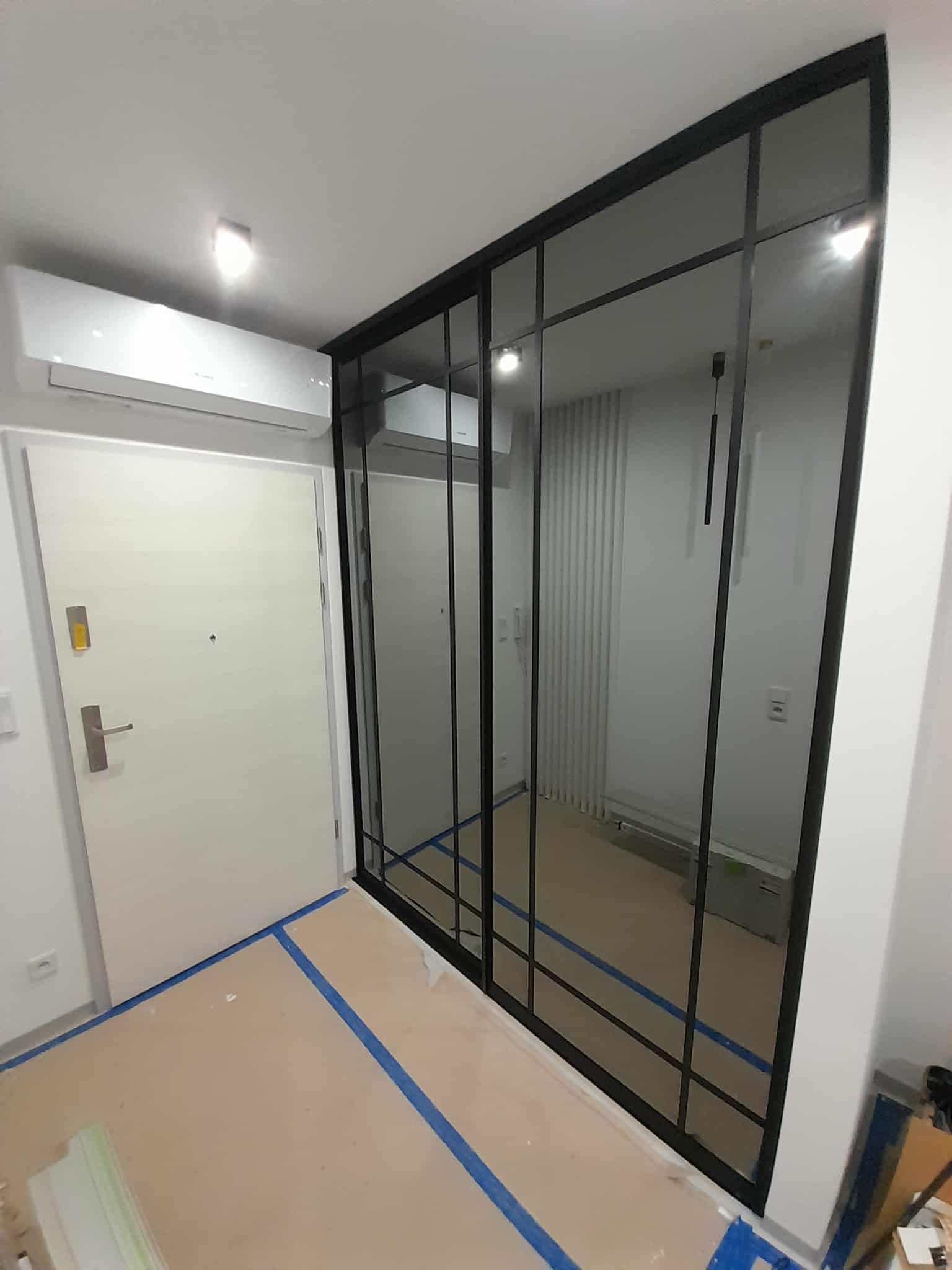 Nowoczesna szafa wnękowa z czarnymi ramami i lustrzanymi drzwiami przesuwnymi, widoczna w korytarzu z zabezpieczoną podłogą i klimatyzacją.