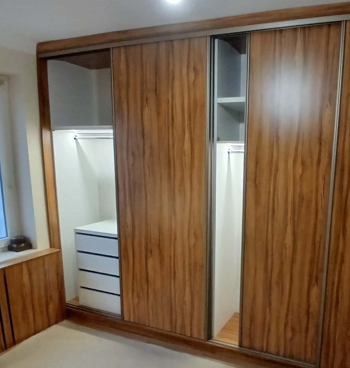 Szafa wnękowa z przesuwnymi drzwiami o drewnianym wzorze, z widocznymi półkami i szufladami wewnątrz. Oświetlenie LED podkreśla wnętrze szafy.