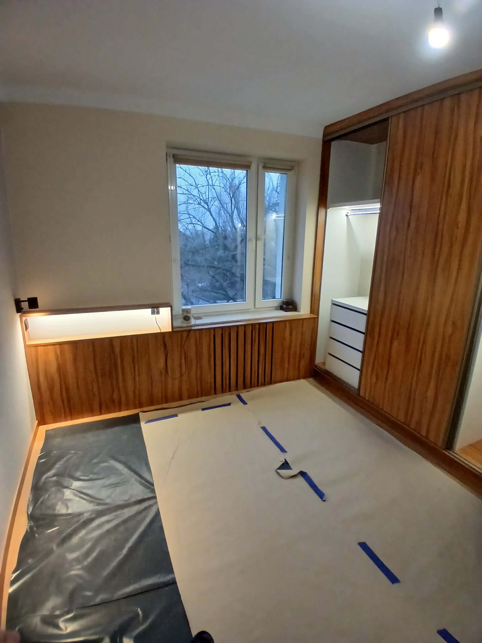 Montaż szafy wnękowej z drewnianymi przesuwnymi drzwiami i zabudową pod oknem. Podłoga zabezpieczona folią i kartonem. Widok na okno z drzewami.