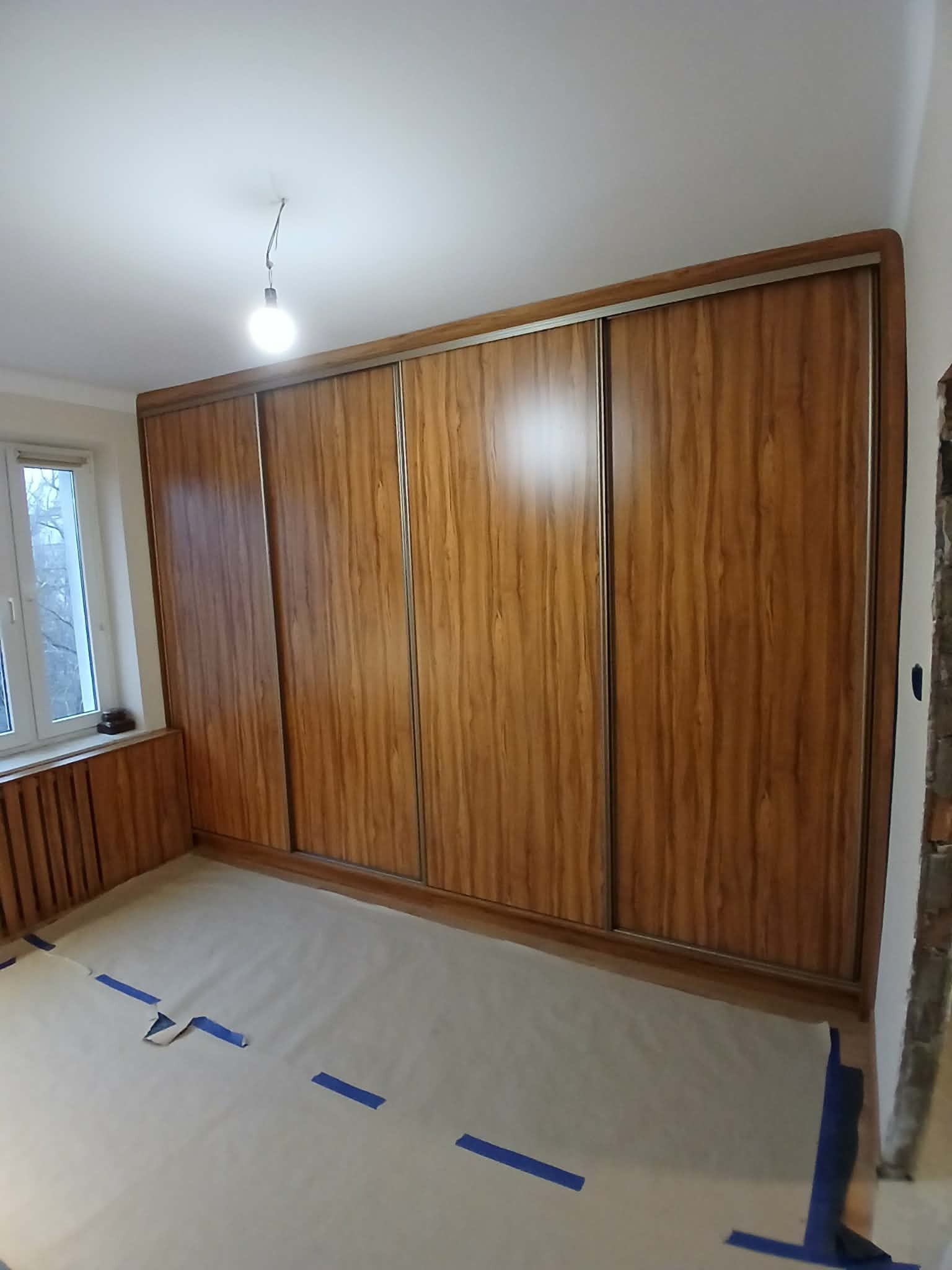 Drewniana szafa wnękowa z przesuwnymi drzwiami, widoczna w pokoju z zabezpieczoną podłogą. Ujęcie z perspektywy podłogi, z widocznym oknem po lewej stronie.