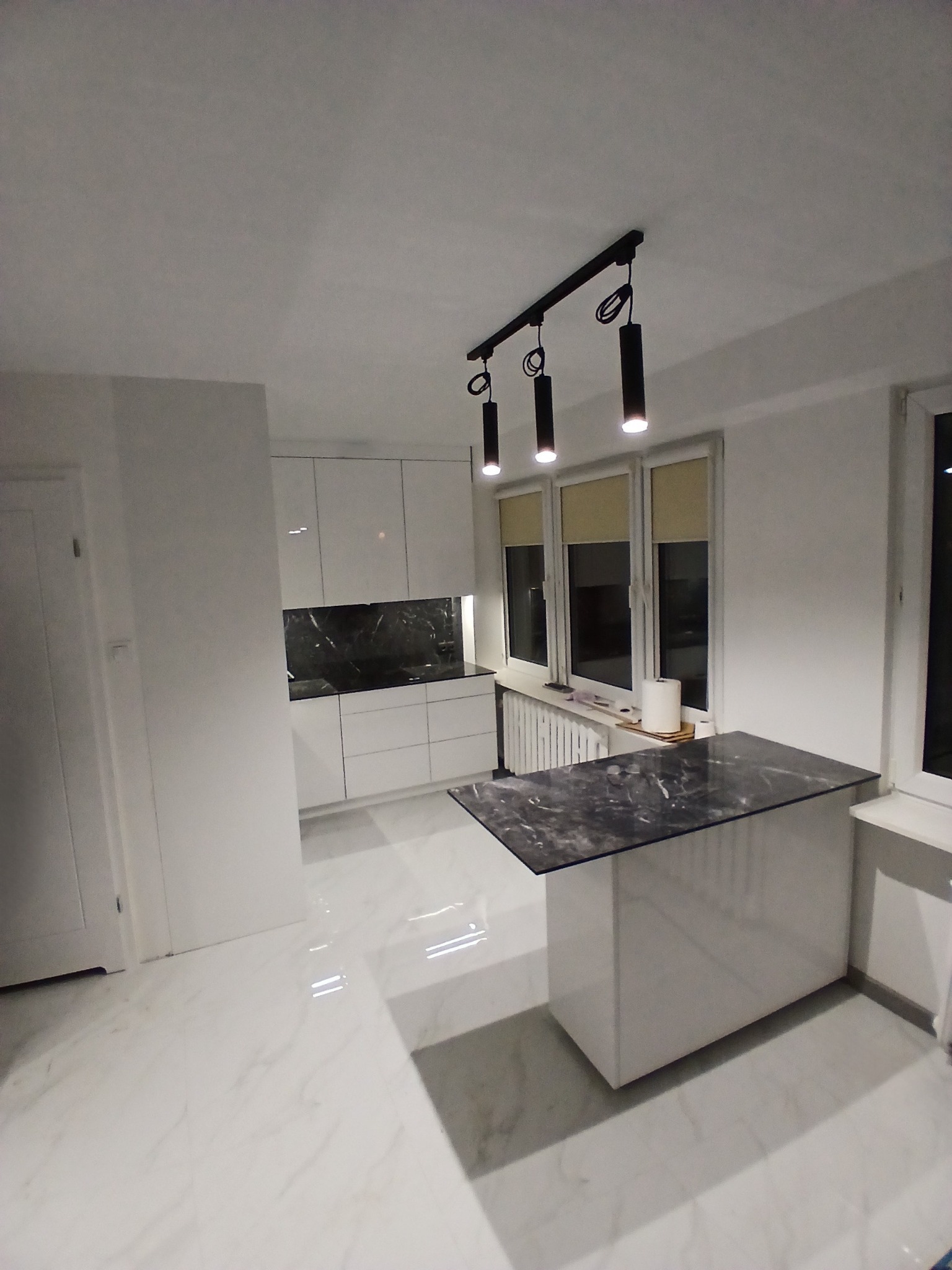 Nowoczesna, biała kuchnia z wyspą o marmurowym blacie, oświetlona trzema reflektorami. Błyszczące fronty szafek i marmurowa podłoga dodają elegancji.