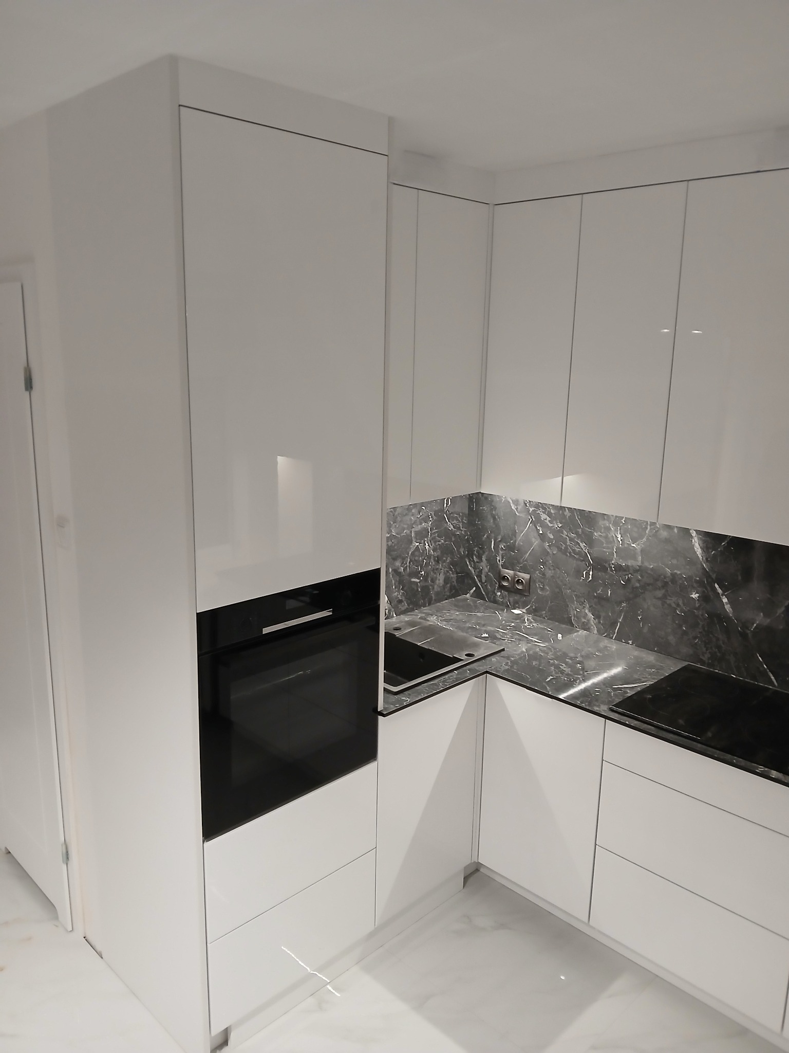 Biała, nowoczesna kuchnia z połyskiem, czarnym blatem imitującym marmur i wbudowanym piekarnikiem. Minimalistyczny design, jasne wnętrze.