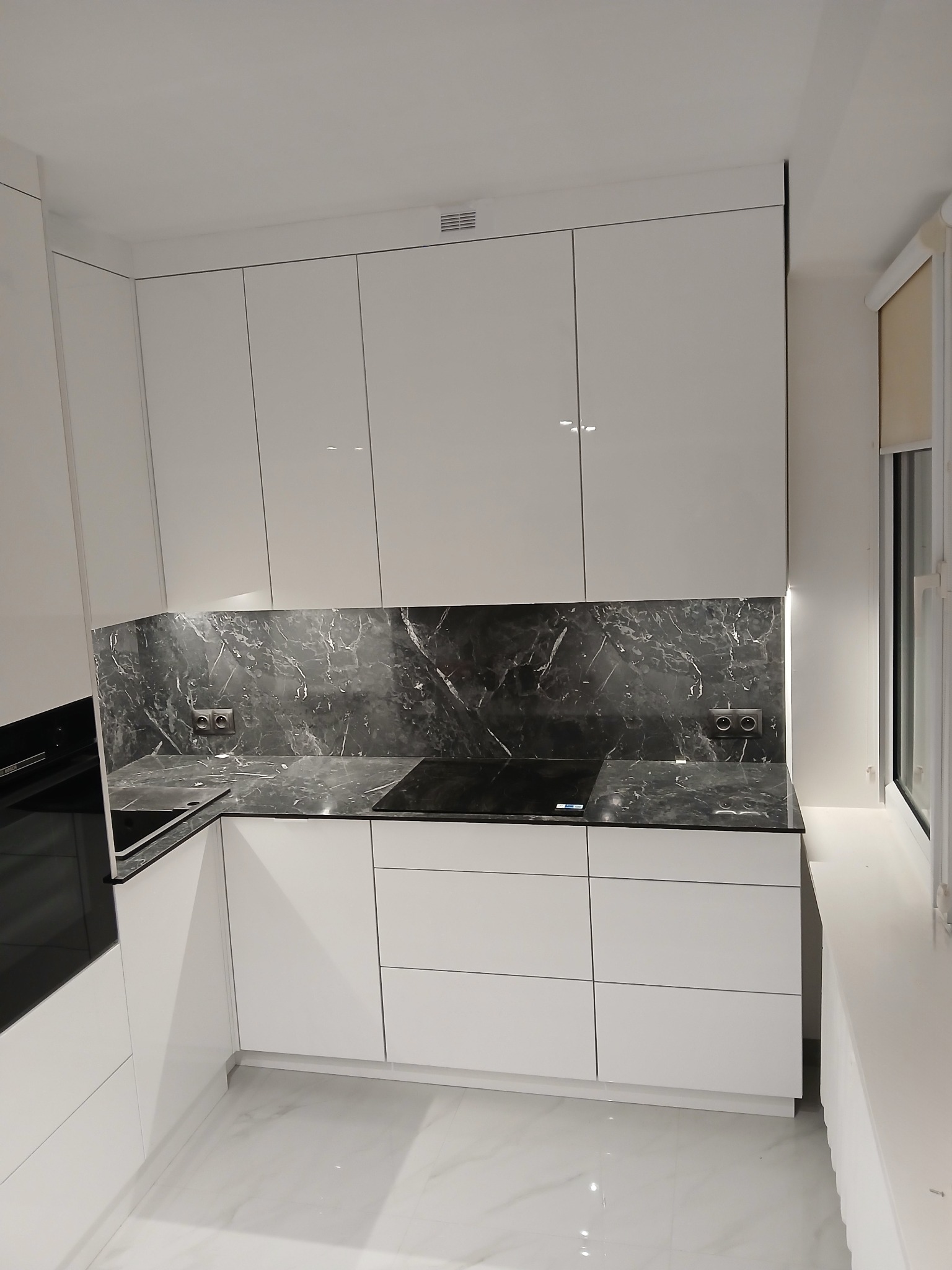 Biała, nowoczesna kuchnia na wymiar z marmurowym blatem i frontami na wysoki połysk. Widoczne gniazdka elektryczne i płyta indukcyjna. Minimalistyczny design.