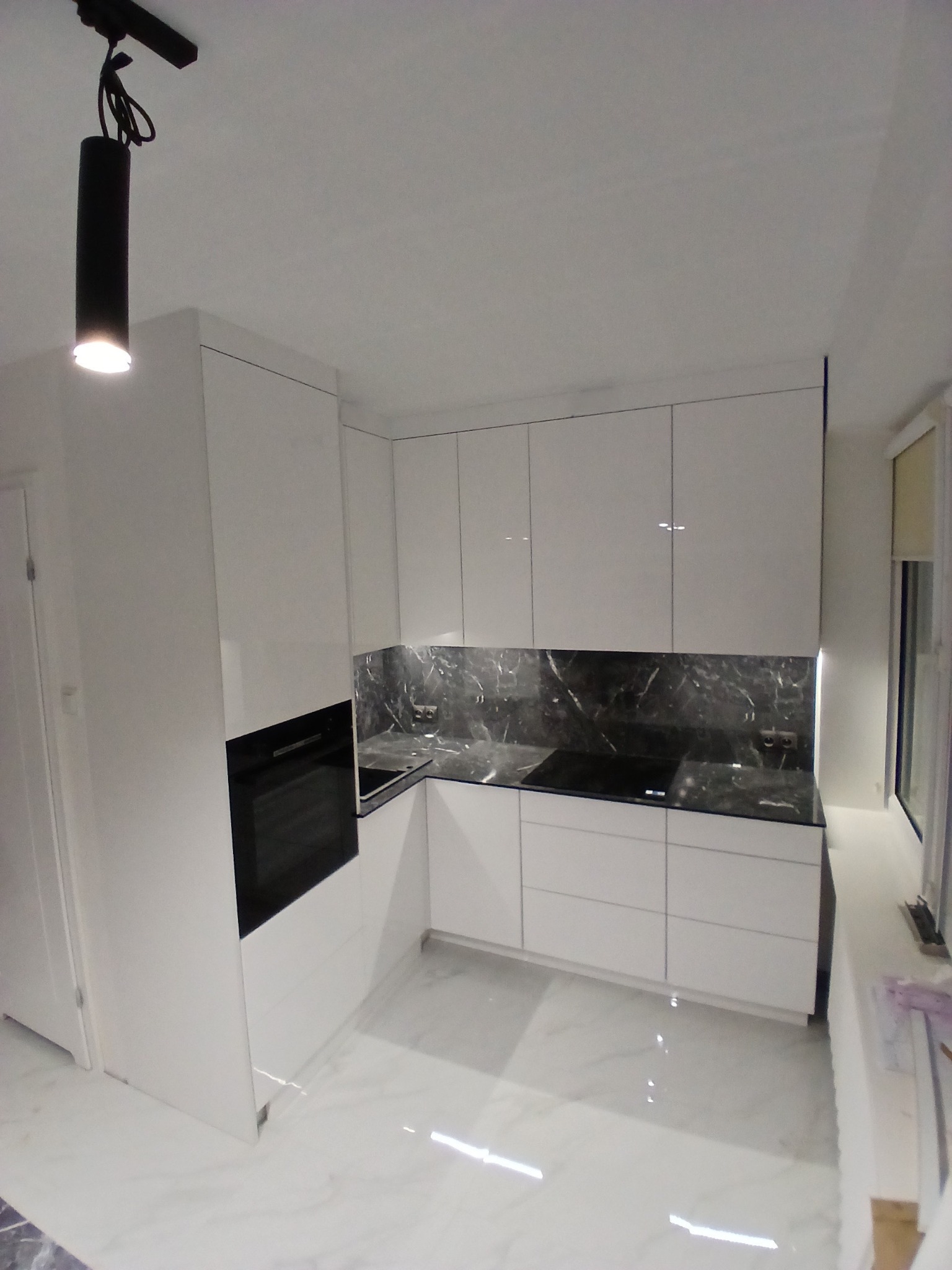 Biała, nowoczesna kuchnia z marmurowym blatem i czarnym oświetleniem punktowym. Minimalistyczny design, błyszczące fronty szafek i płyta indukcyjna.