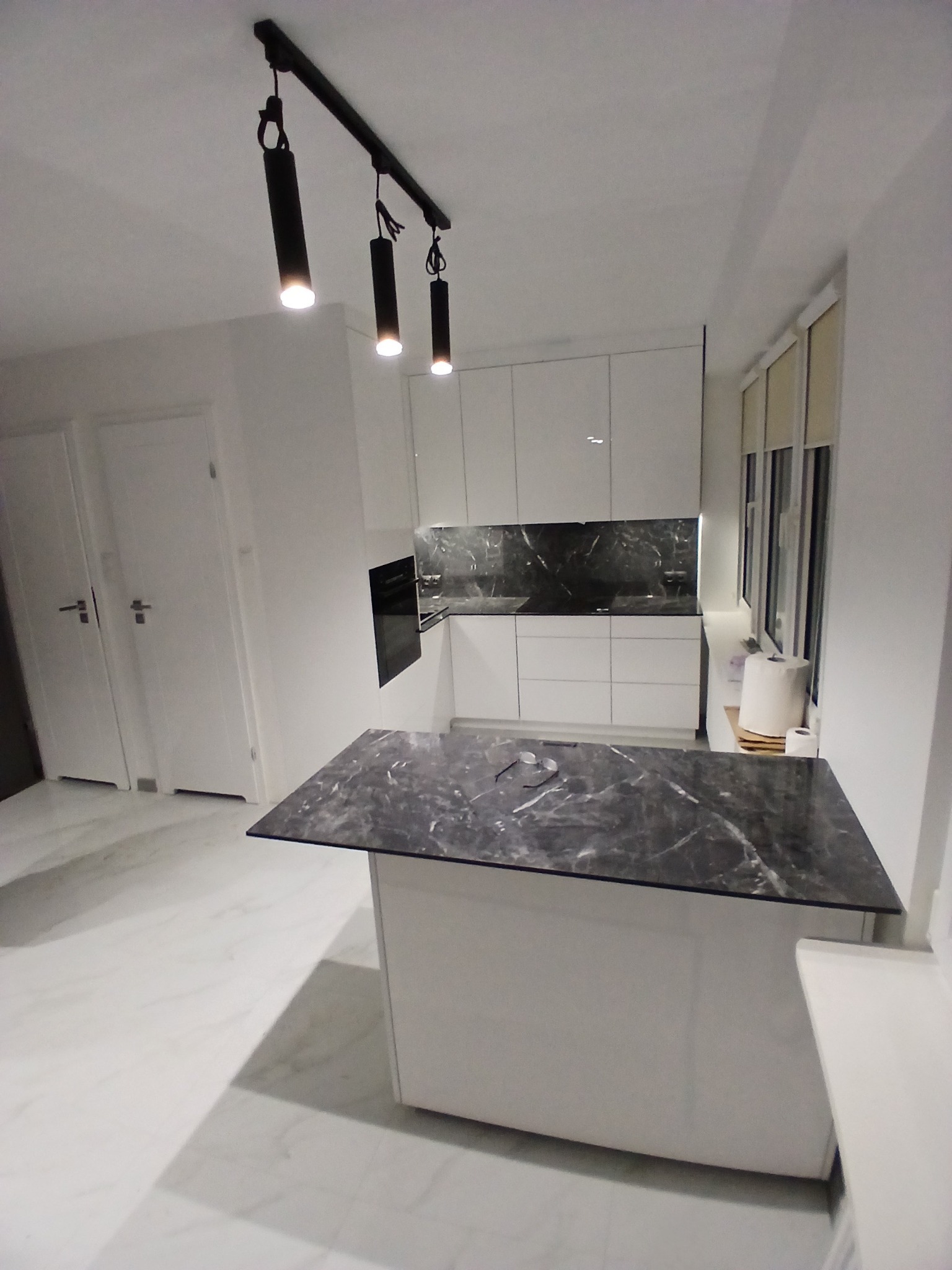 Nowoczesna, biała kuchnia z wyspą i blatem imitującym marmur, oświetlona czarnymi lampami punktowymi. Minimalistyczny design, białe fronty szafek, marmurowy backsplash.