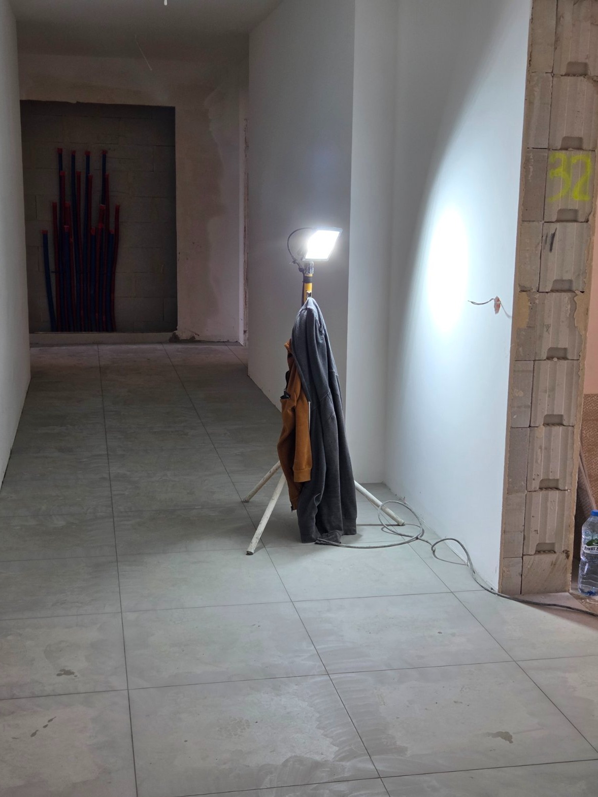 Wykończone płytki podłogowe w korytarzu w trakcie remontu, oświetlone lampą na statywie. W tle widoczne rury instalacyjne w ścianie.