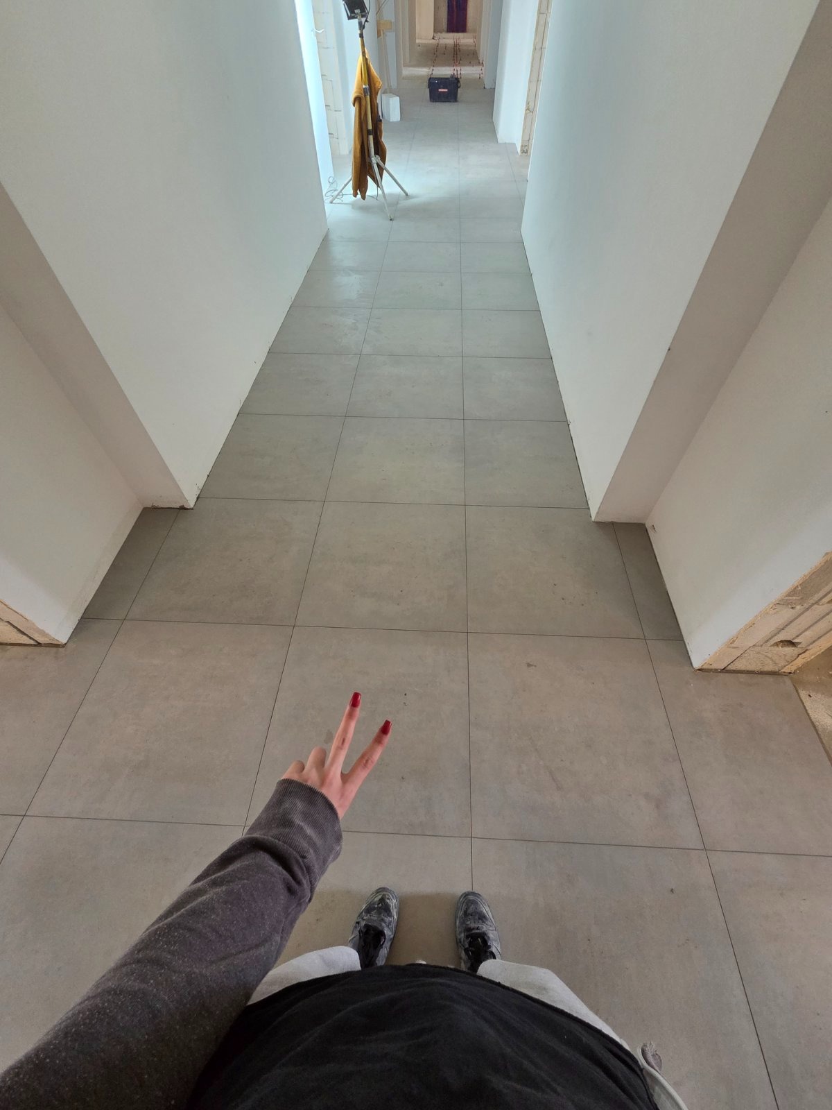 Długi korytarz z szarymi płytkami podłogowymi, oświetlony lampą. W tle widoczny sprzęt. Na pierwszym planie dłoń z czerwonymi paznokciami i fragment osoby.