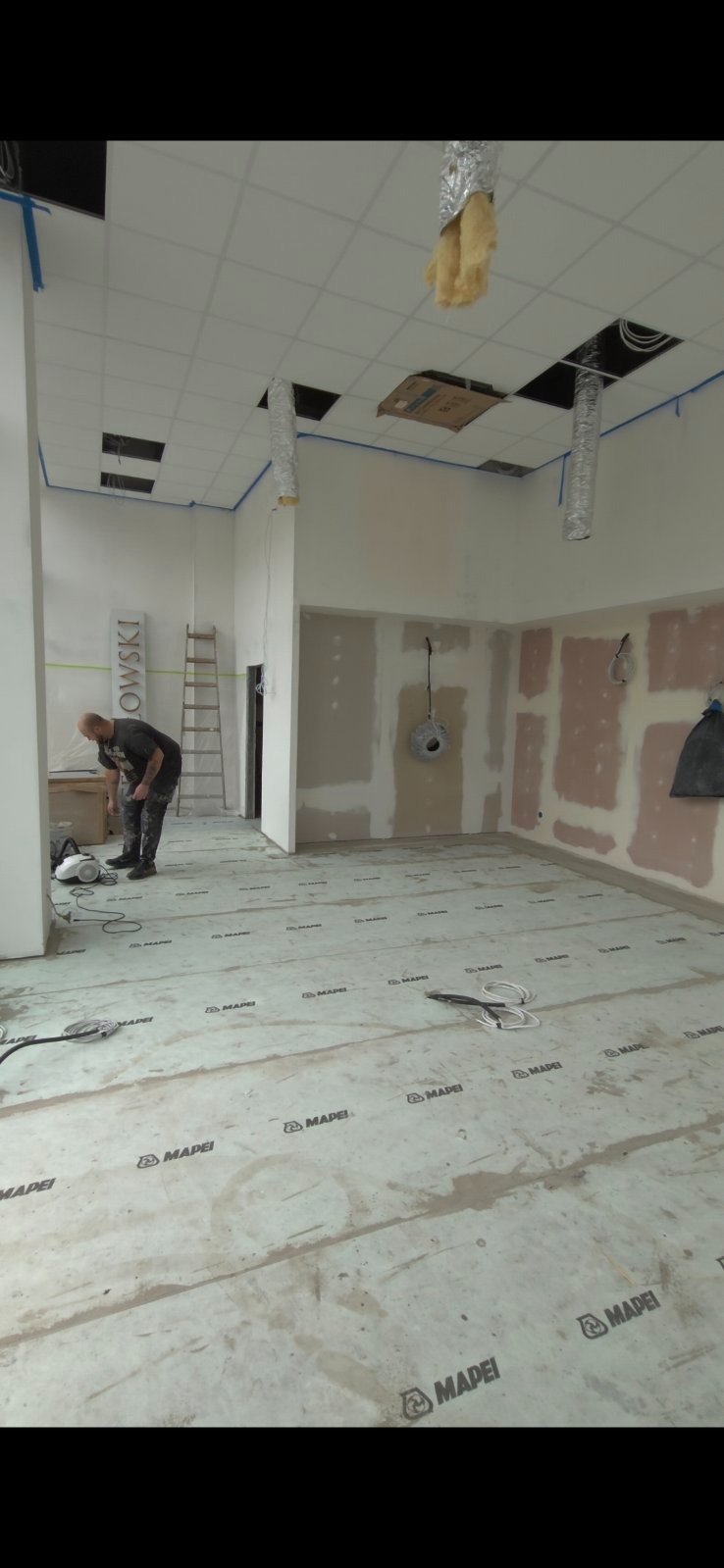 Wnętrze w trakcie prac wykończeniowych: podłoga zabezpieczona folią Mapei, ściany częściowo wygładzone, widoczny pracownik i instalacje sufitowe.