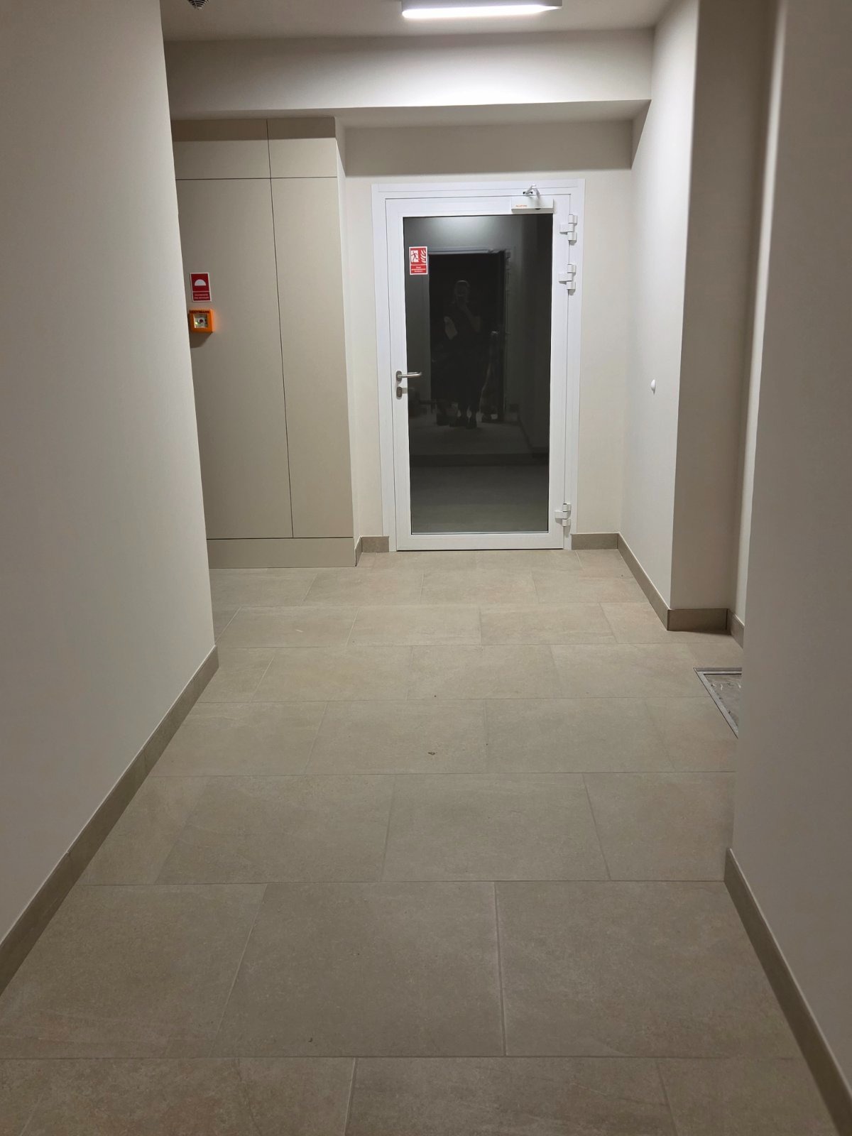 Beżowy korytarz z płytkami podłogowymi w neutralnych odcieniach. Białe ściany i drzwi wejściowe. Widoczny fragment instalacji przeciwpożarowej.