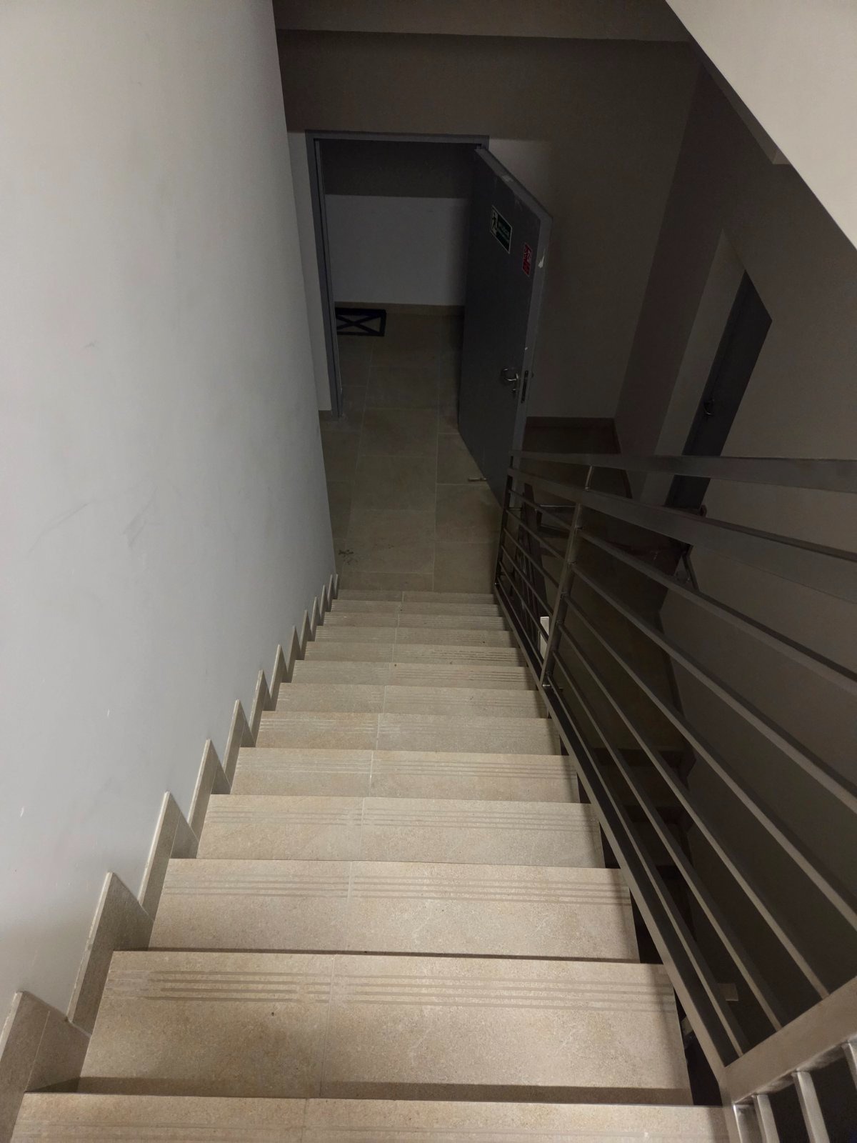 Klatka schodowa z jasnymi, wyłożonymi płytkami schodami i metalową barierką. W tle widoczne drzwi ewakuacyjne i fragment posadzki.