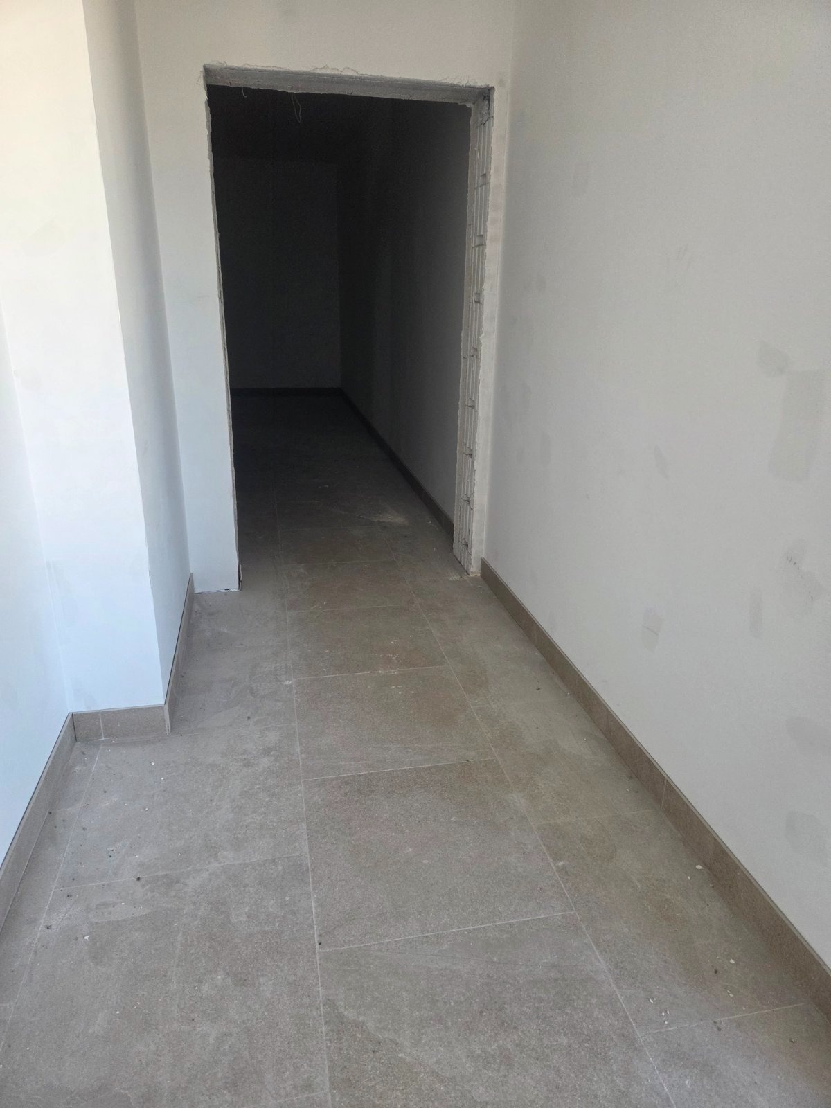 Wykończony korytarz z szarymi płytkami podłogowymi i świeżo pomalowanymi ścianami na biało. Widoczne surowe wykończenie ościeżnicy.