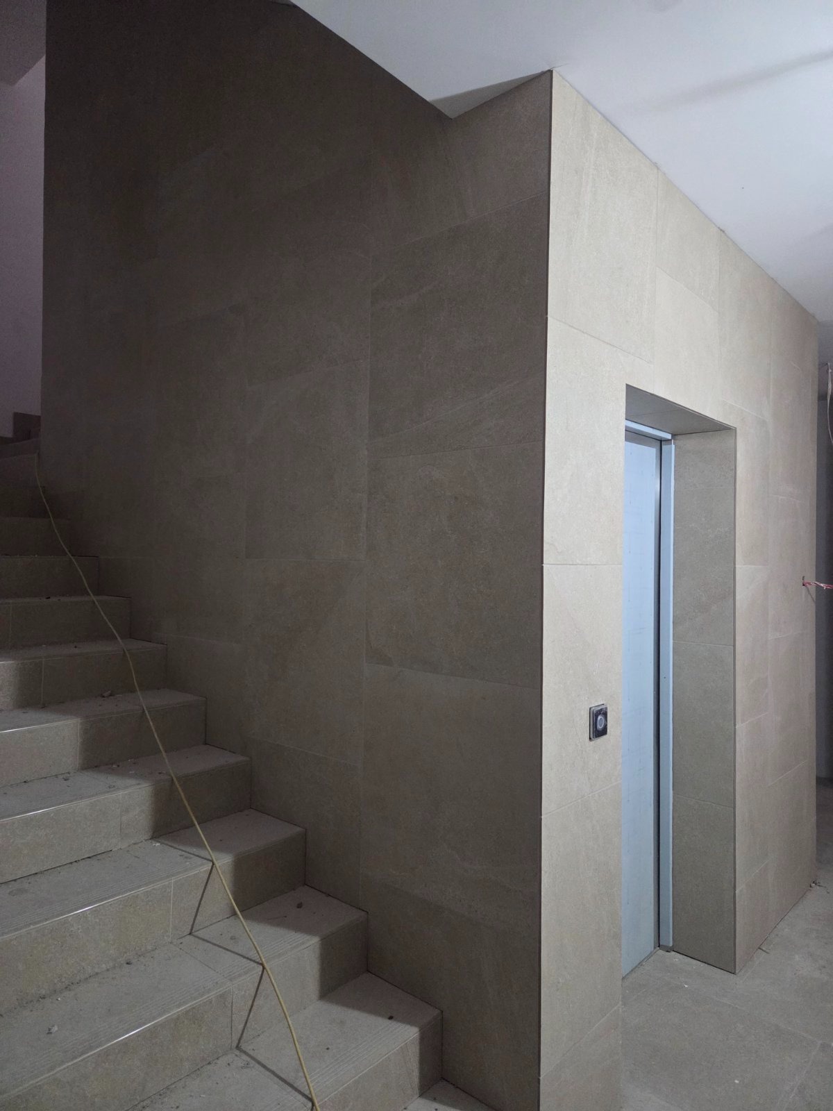 Wykończenie klatki schodowej beżowymi płytkami imitującymi kamień. Widoczne schody, ściany w płytkach i obudowa drzwi. Minimalistyczny design.