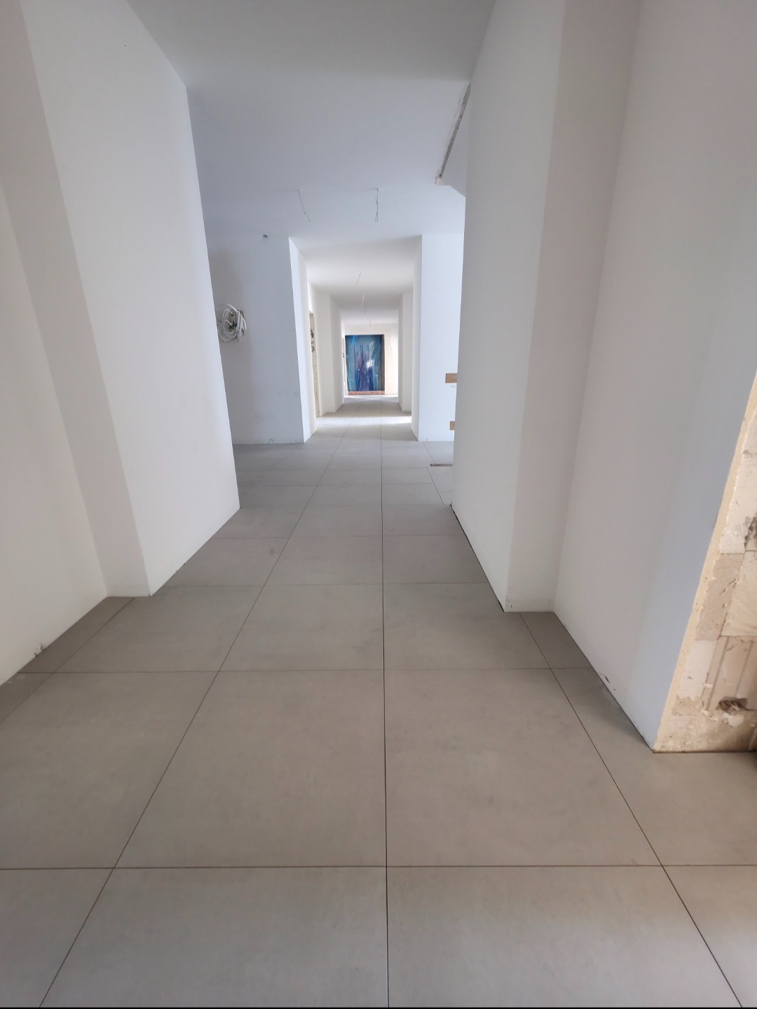 Długi korytarz z szarymi płytkami podłogowymi i białymi ścianami. W oddali widoczne niebieskie obrazy. Perspektywa zbieżna, podkreślająca głębię.