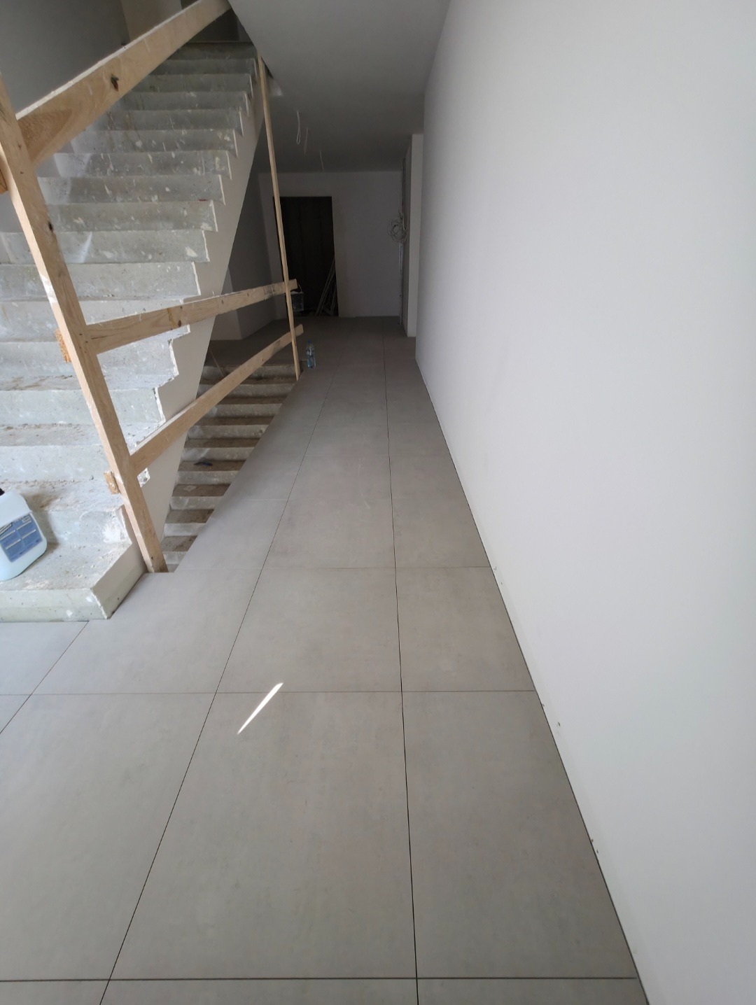 Korytarz w trakcie wykańczania: ułożona jasna terakota, surowe schody z drewnianą barierką, białe ściany. Widoczny fragment pojemnika z klejem do płytek.