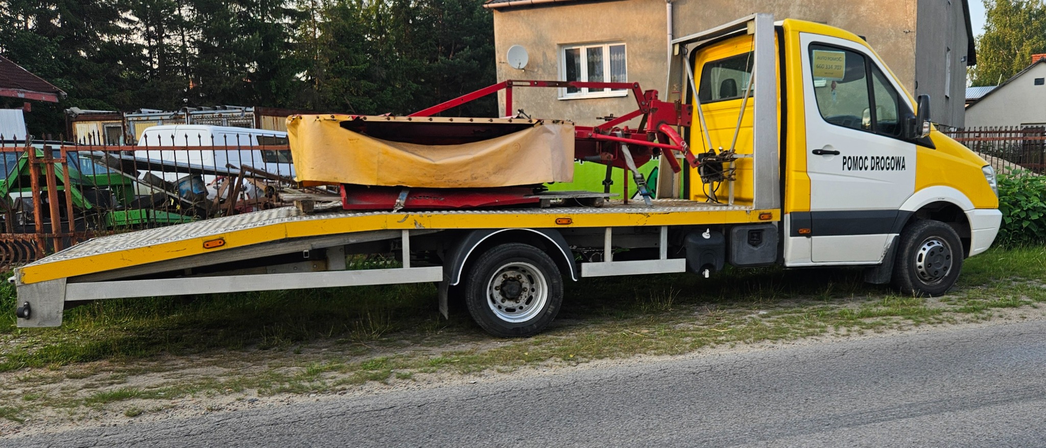 Żółto-biała laweta z napisem 'Pomoc Drogowa' przewozi maszynę rolniczą na platformie. Widoczne drzewa i budynek w tle. Słoneczny dzień.