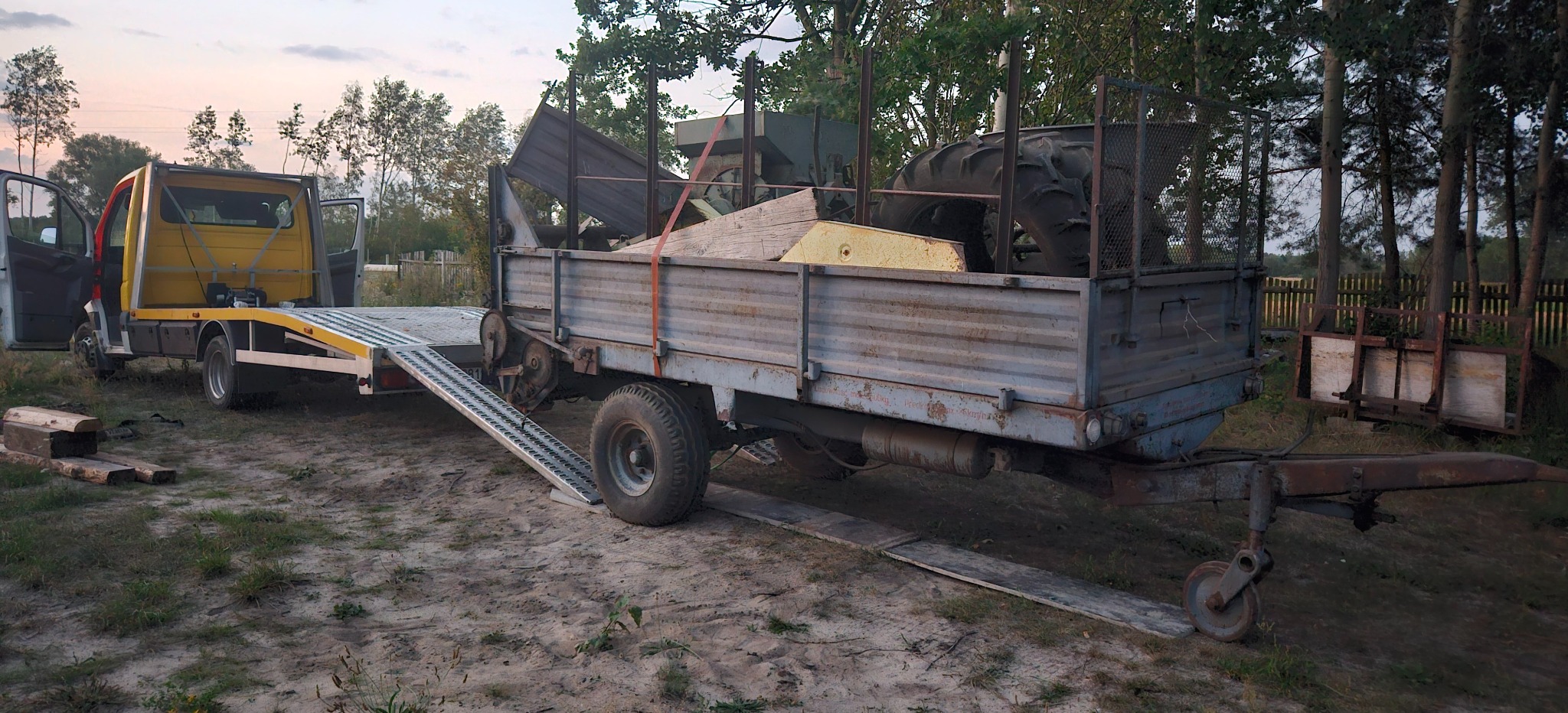 Żółta laweta z rozłożoną rampą i przyczepą wypełnioną ładunkiem (m.in. koło od traktora) na piaszczystym podłożu, w tle drzewa i zachmurzone niebo.