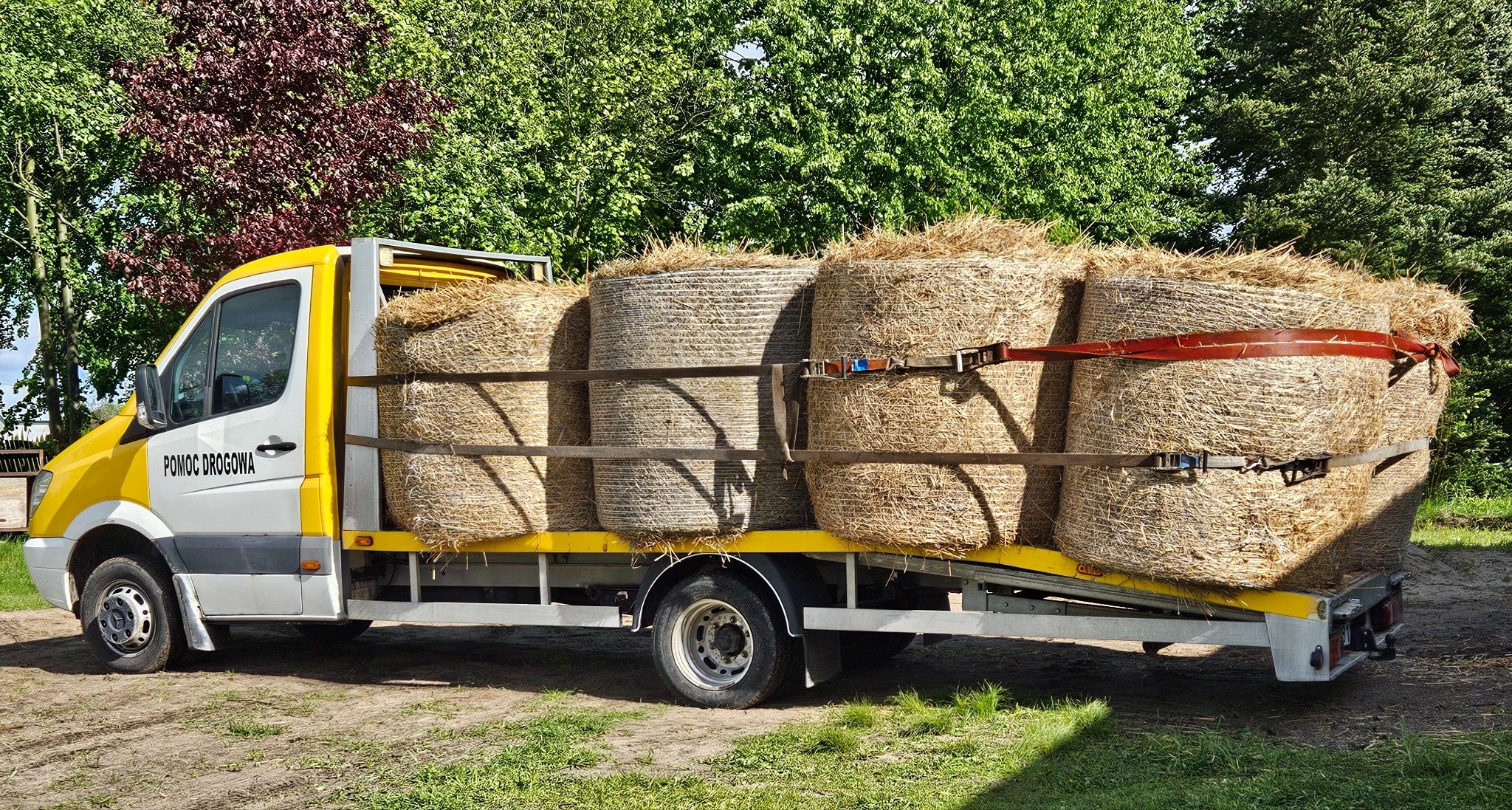 Żółto-biała laweta z napisem 'Pomoc Drogowa' przewozi bele siana, zabezpieczone pasami. Tło stanowią zielone drzewa.