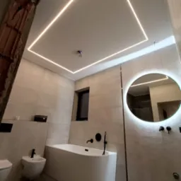 Nowoczesna łazienka z podświetlanym sufitem, okrągłym lustrem z ledami i minimalistyczną wanną. Widoczny fragment ściany w trakcie prac wykończeniowych.