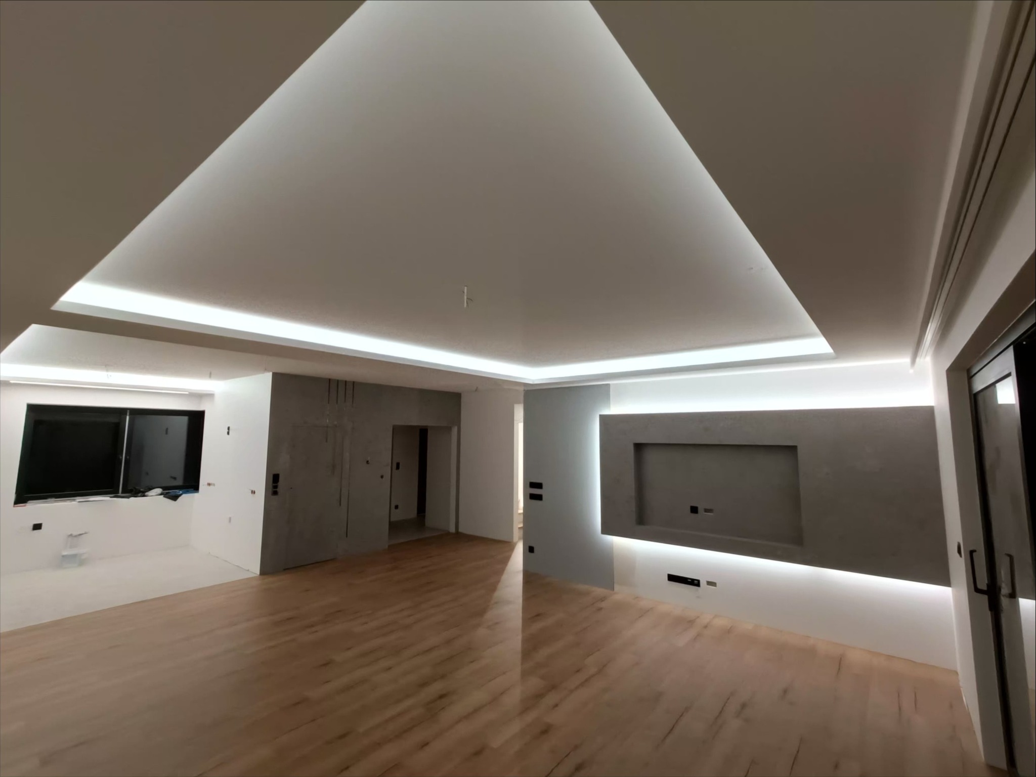 Nowoczesne wnętrze po remoncie: drewniana podłoga, szara ściana z wnęką na TV, podświetlany sufit. Minimalistyczny design, świeżo po wykończeniu.