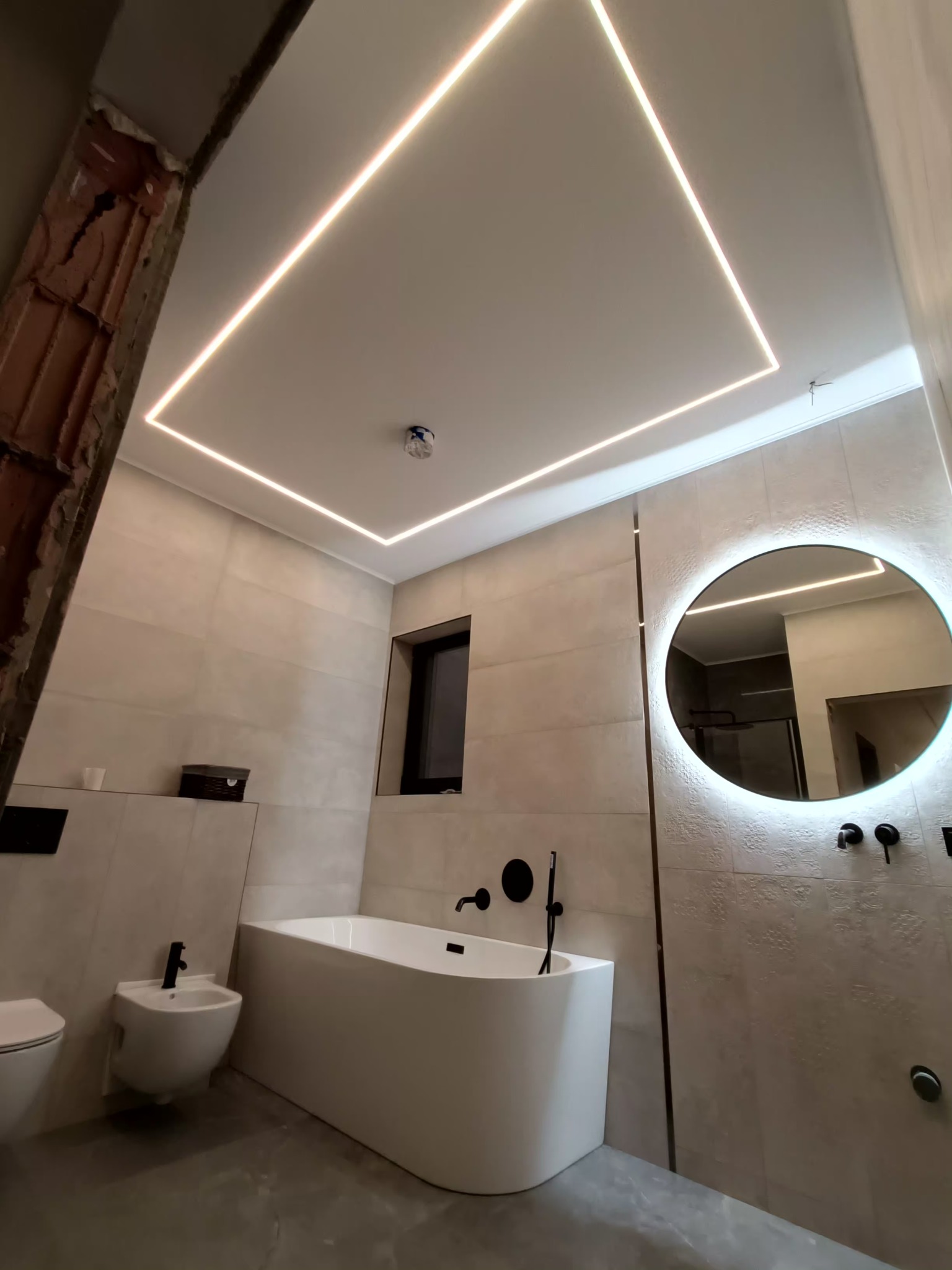Nowoczesna łazienka z podświetlanym sufitem, okrągłym lustrem z ledami i minimalistyczną wanną. Widoczny fragment ściany w trakcie prac wykończeniowych.
