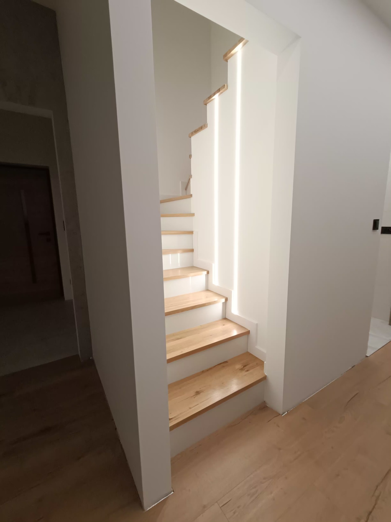 Nowoczesne schody z drewnianymi stopniami i białymi podstopnicami, oświetlone pionowymi pasami światła, w minimalistycznym wnętrzu.