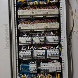 Rozdzielnica elektryczna z licznymi bezpiecznikami, przewodami oznaczonymi etykietami i elementami sterującymi, zamontowana w metalowej obudowie marki Legrand.