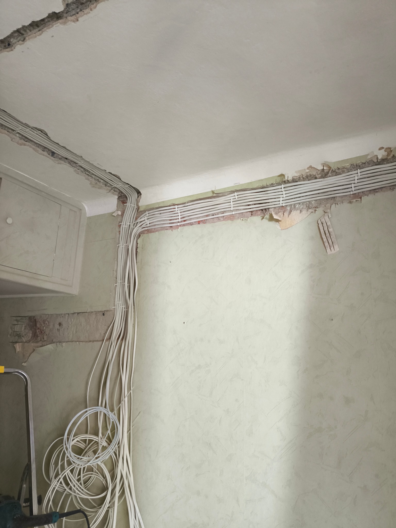 Białe kable elektryczne ułożone wzdłuż ściany i sufitu w trakcie remontu. Widoczne uchwyty mocujące i pozostałości tapety. Częściowo ułożone przewody leżą na podłodze.
