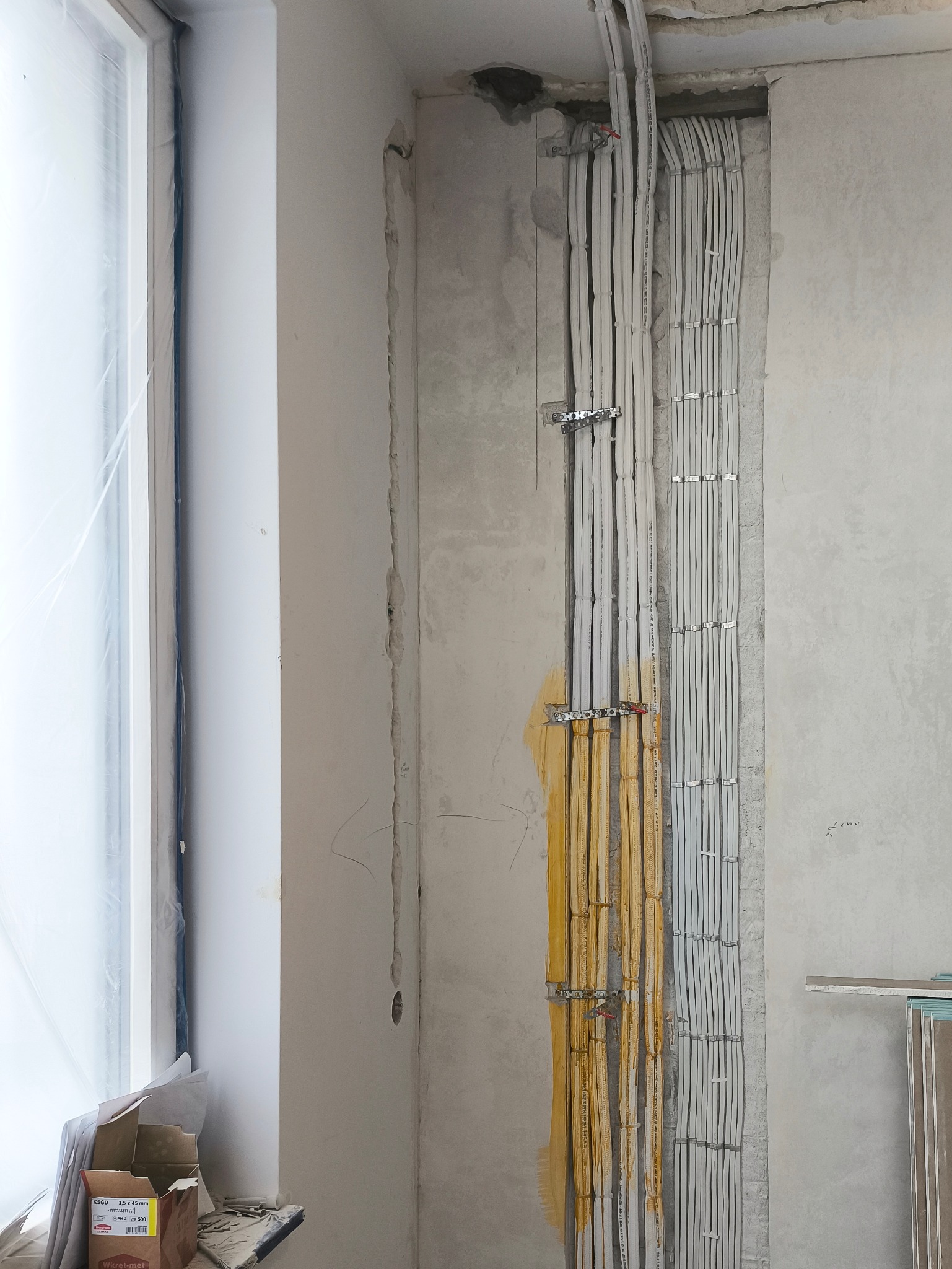 Ściana w trakcie montażu instalacji elektrycznej w Łodzi. Widoczne kable zabezpieczone metalowymi uchwytami, surowy beton i okno z folią ochronną.