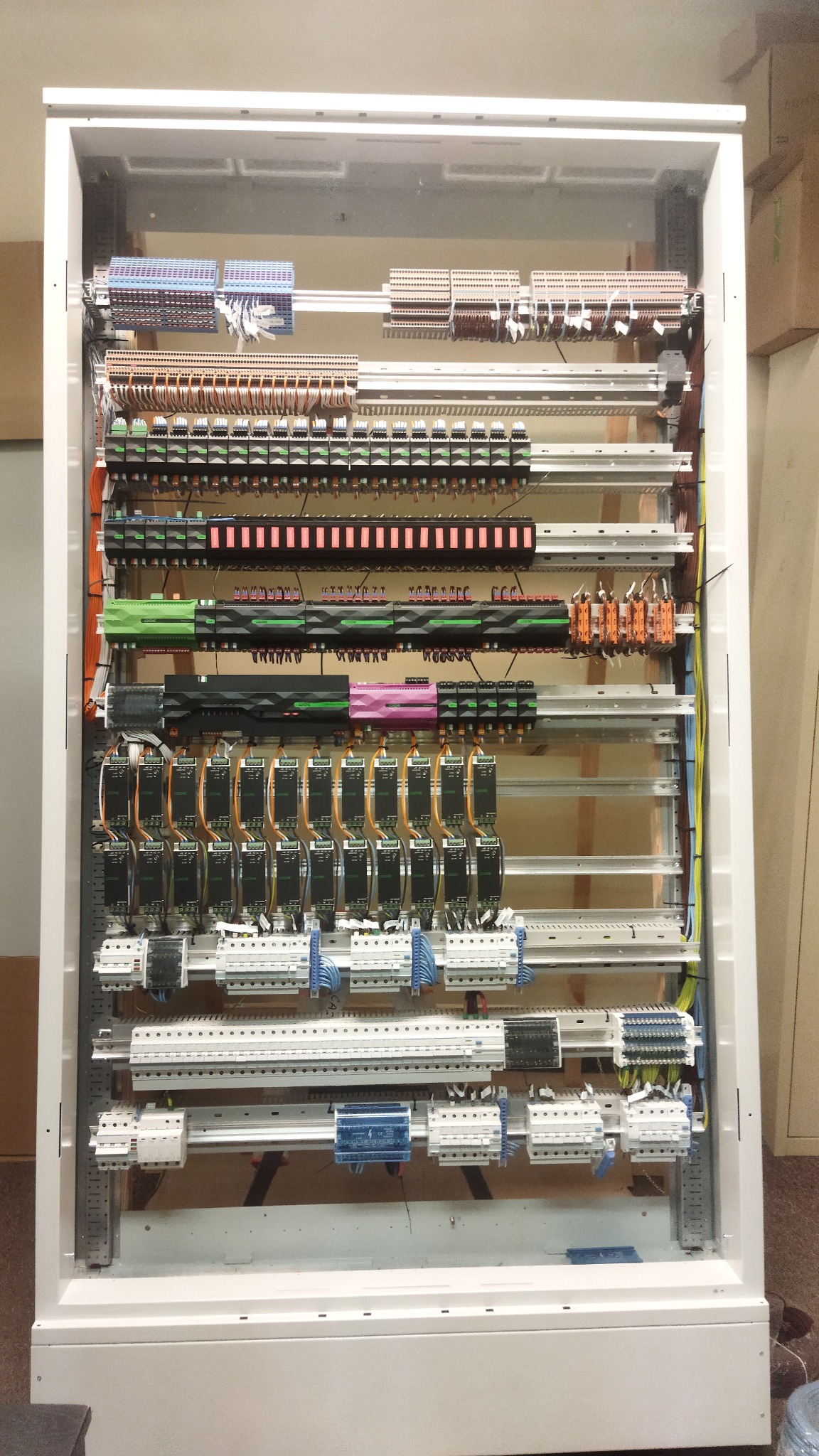 Rozdzielnica elektryczna w białej szafie, widoczne liczne przewody, listwy zaciskowe i moduły sterujące. Ułożone poziomo, równolegle do siebie, uporządkowane okablowanie.