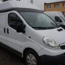 Biały Opel Vivaro z wysokim dachem na parkingu, widok z przodu. W tle budynek i inny bus. Lekko pochmurna pogoda.