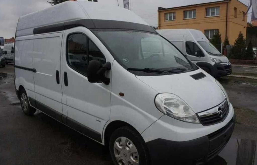 Biały Opel Vivaro z wysokim dachem na parkingu, widok z przodu. W tle budynek i inny bus. Lekko pochmurna pogoda.