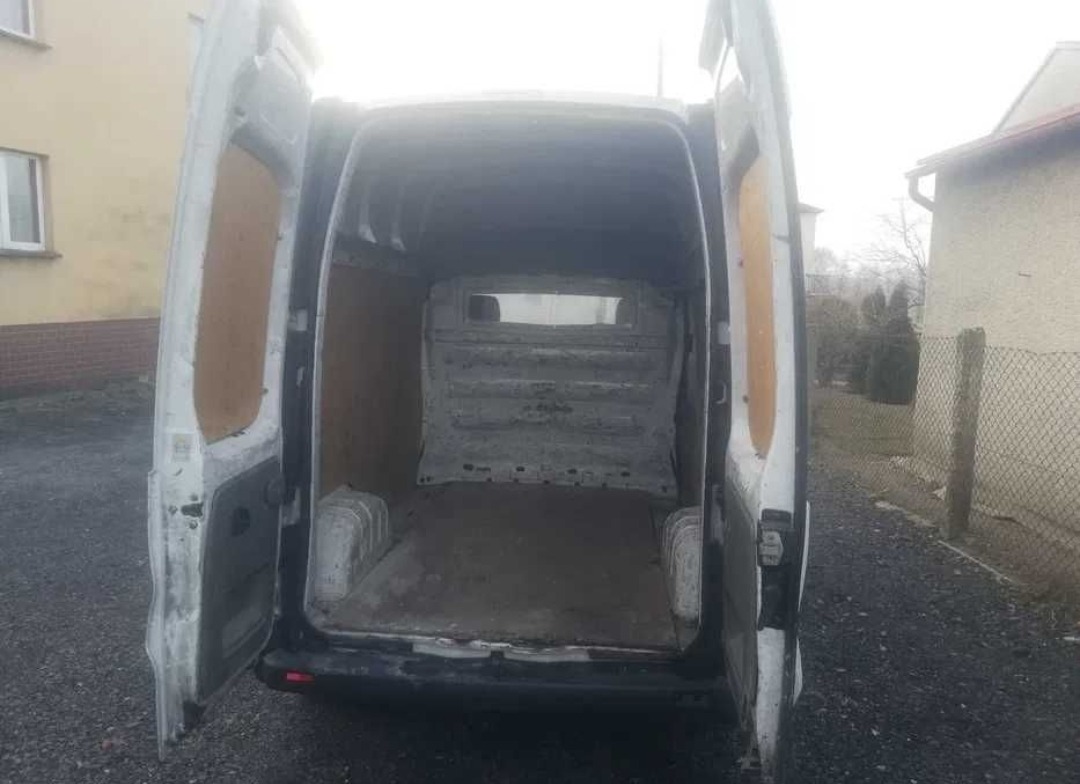 Wnętrze białego, pustego furgonetki z otwartymi drzwiami, widok z tyłu na przestrzeń ładunkową. Widoczne ślady użytkowania.