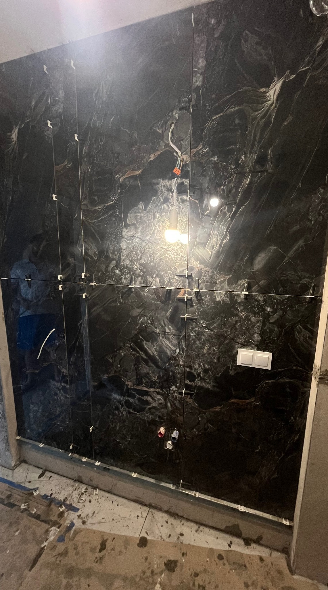 Ściana wyłożona czarnymi, marmurkowymi płytkami z białymi krzyżykami dystansowymi, widoczne przewody elektryczne i włącznik. W tle odbicie osoby.