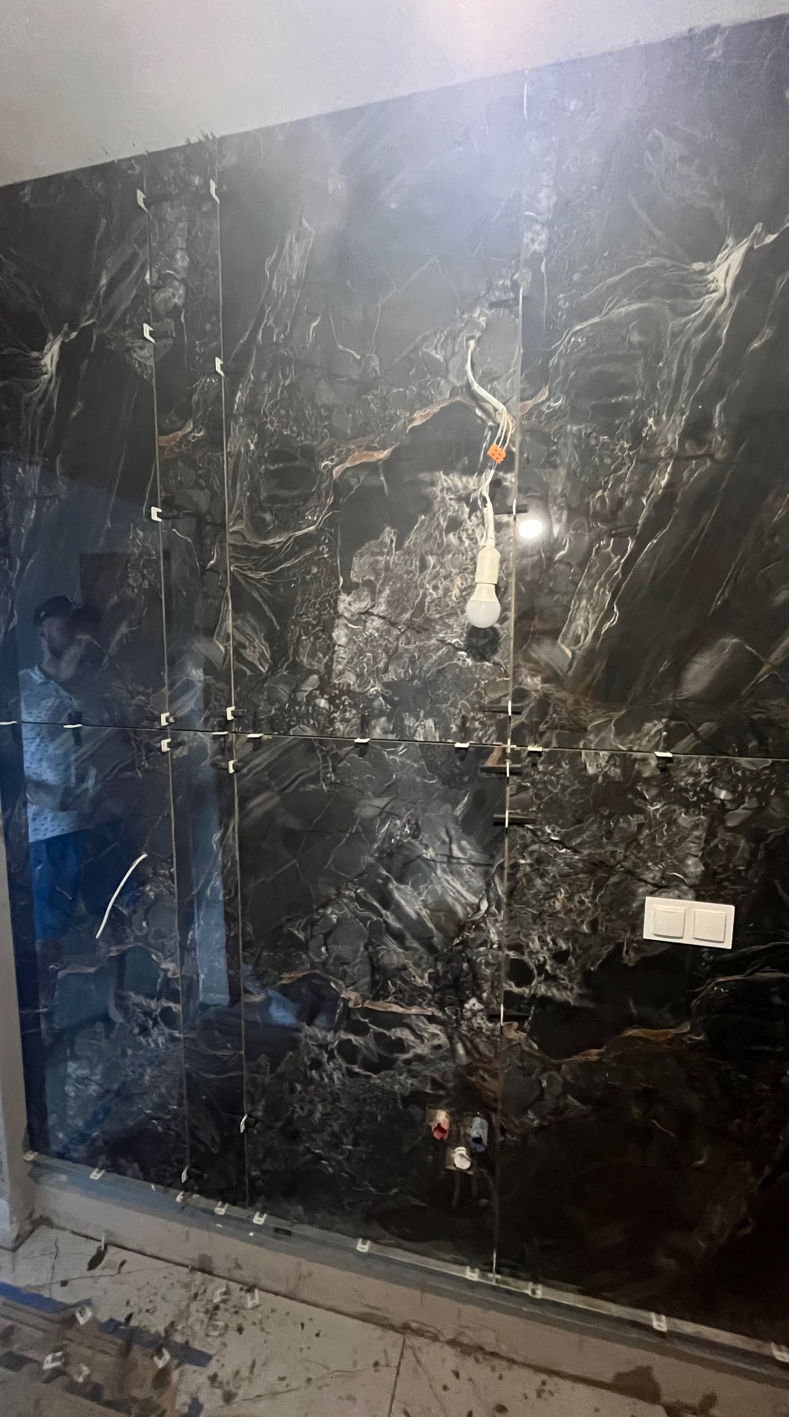 Częściowo wykończona ściana z czarnych, marmurkowych płytek w łazience. Widoczne krzyżyki dystansowe, instalacja elektryczna i fragment podłogi z listwą.
