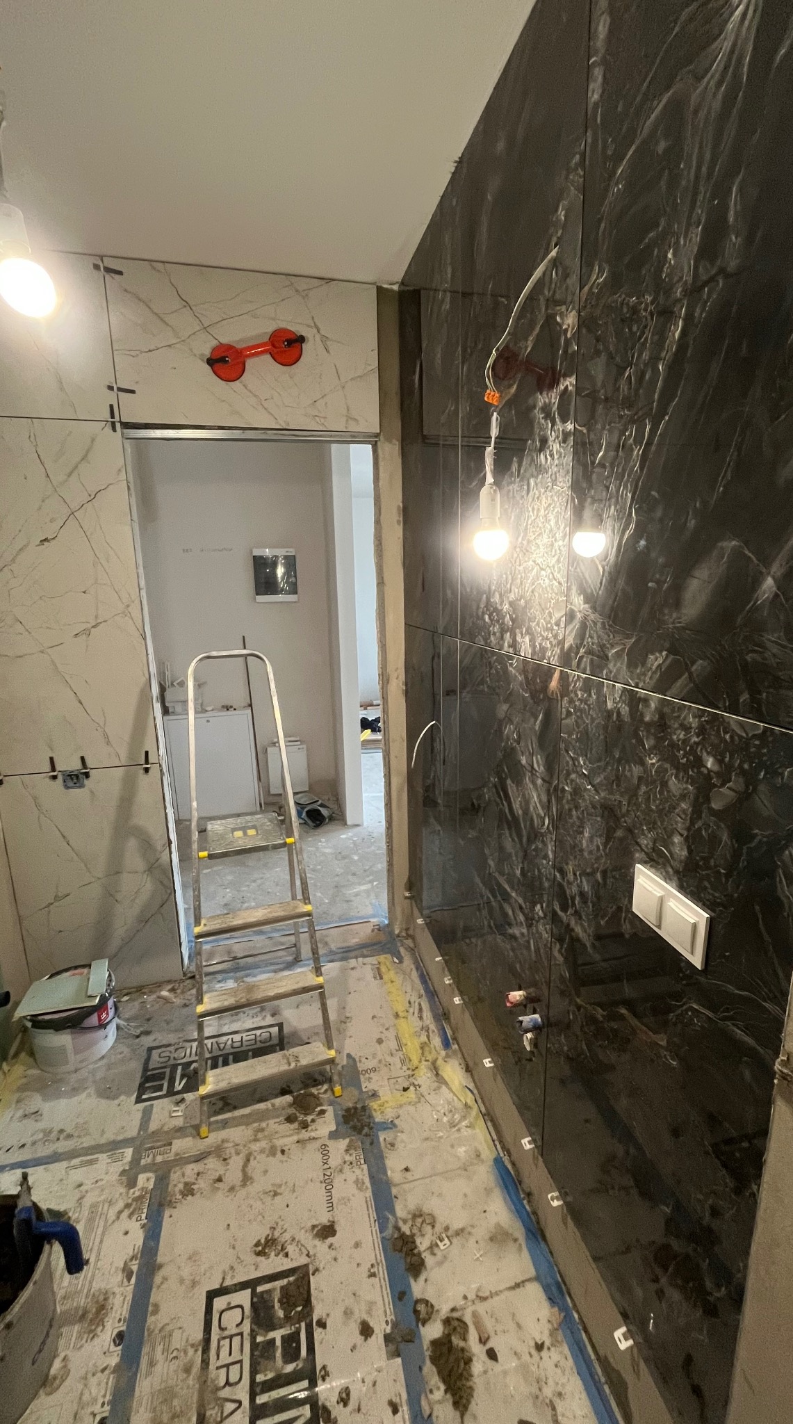 Montaż płytek w łazience: czarne marmurowe płytki na ścianie, drabina, przyssawki do płytek, białe płytki imitujące marmur w tle. Wnętrze w trakcie wykończenia.