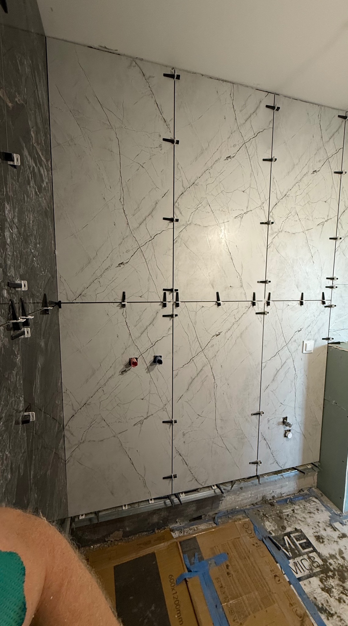 Montaż płytek imitujących marmur na ścianie z użyciem klipsów poziomujących. Widoczne fragmenty podłogi i ściany obok. Częściowo widoczna noga glazurnika.