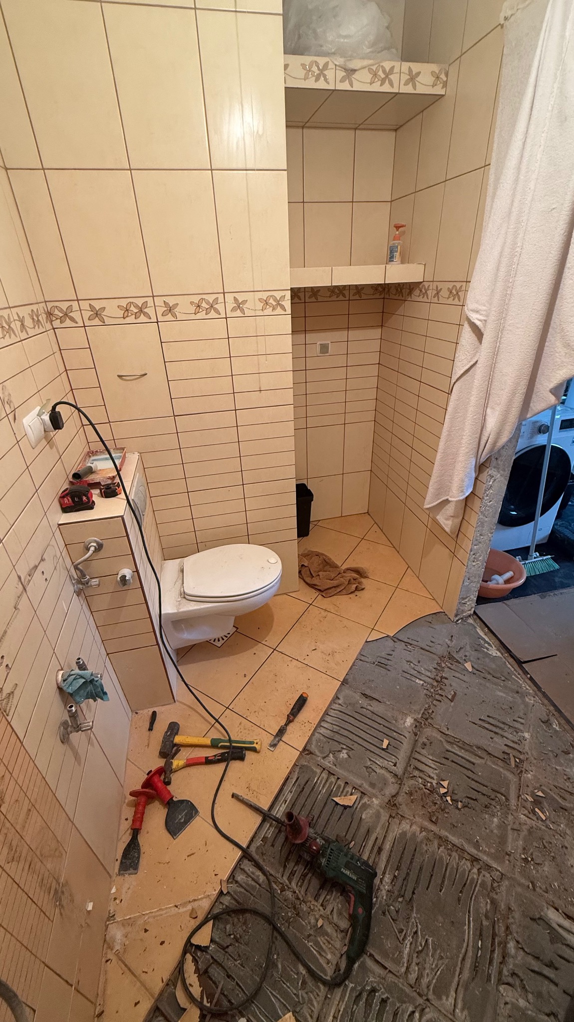 Demontaż płytek w łazience z beżowymi kaflami. Widoczne narzędzia: wiertarka, młotki, szpachelki. Podłoga częściowo bez płytek, z widocznymi śladami po kleju.