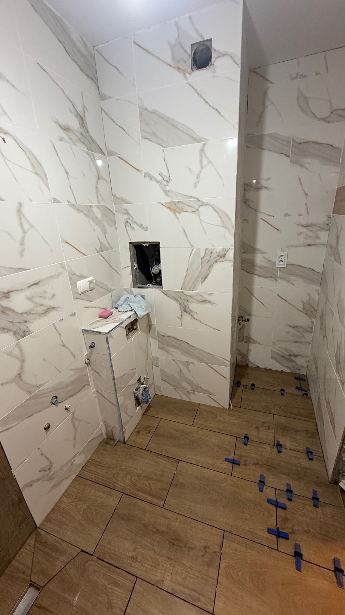Łazienka w trakcie wykańczania: marmurowe płytki na ścianach, drewnopodobne na podłodze, niebieskie krzyżyki dystansowe, zabudowa stelaża WC w trakcie prac.