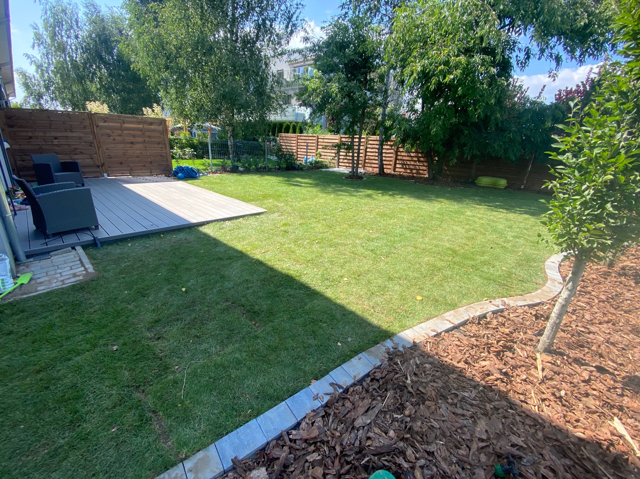 Zadbany ogród z równo przystrzyżonym trawnikiem, drewnianym tarasem i ogrodzeniem. Widoczne drzewa i ozdobna kora wokół jednego z nich. Krzesła ogrodowe na tarasie.