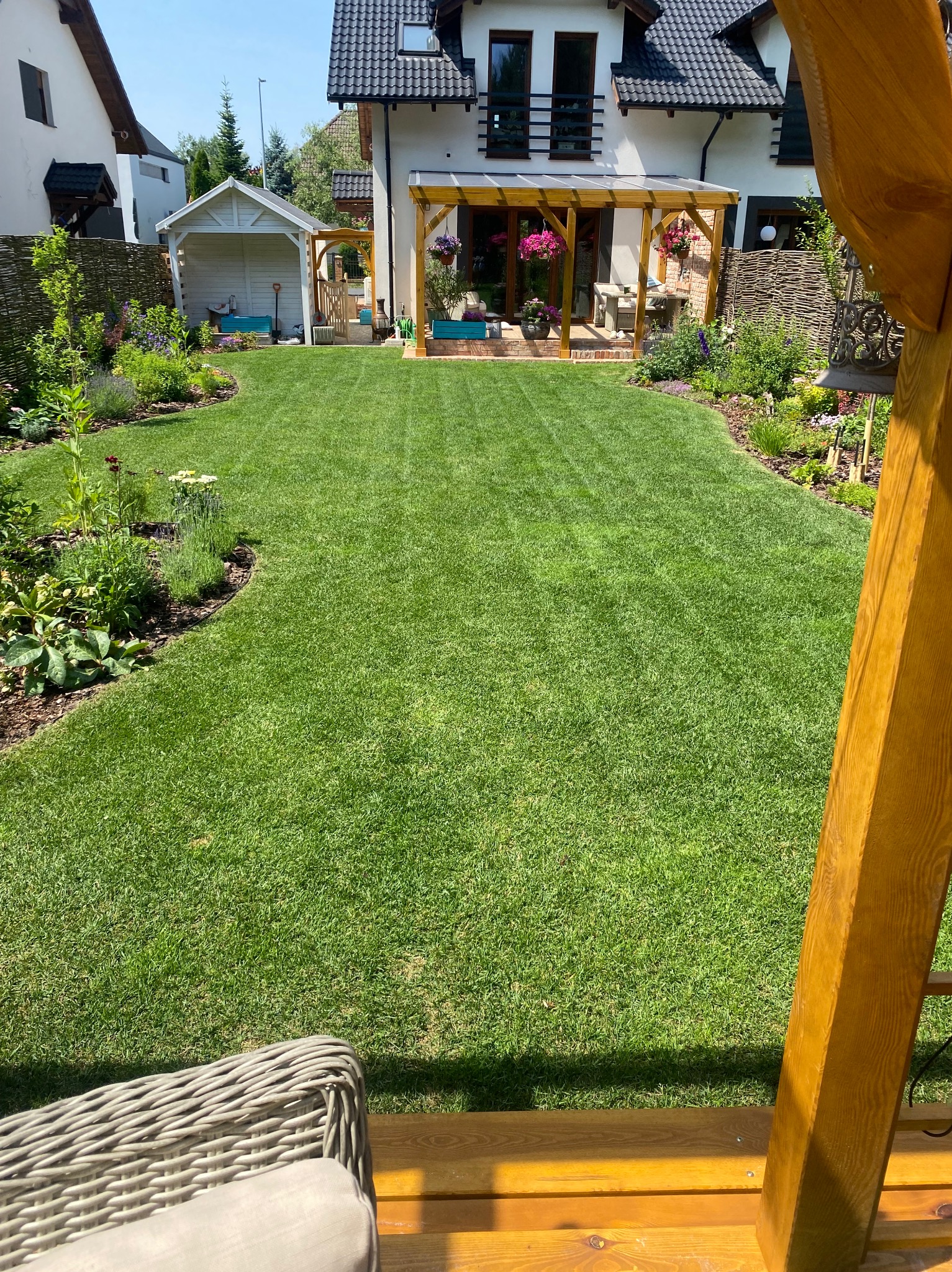Zadbany trawnik z równo przystrzyżoną trawą i rabatami kwiatowymi, widok z huśtawki w kierunku domu z drewnianą pergolą i małym domkiem ogrodowym w tle.