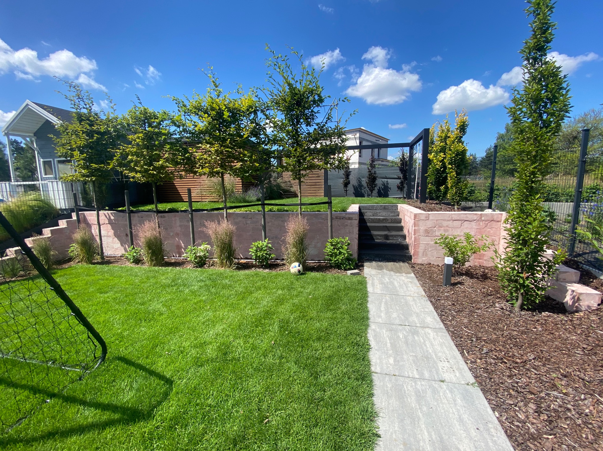 Nowoczesny ogród z trawnikiem, ścieżką z płyt, niskim murem oporowym, nasadzeniami i czarnym ogrodzeniem panelowym. Piękne, słoneczne popołudnie.