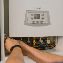 Serwis Fabryczny Urządzeń Gazowo-Elektrycznych - Szymaniak - oddział Szczecin - Instalacja pieca gazowego Termet EcoCondens Gold Plus w szafce. Widoczne zawory i podłączenia. Ręce montera przy pracy, szafka w jasnej kolorystyce.