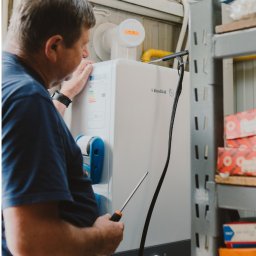 Serwis Fabryczny Urządzeń Gazowo-Elektrycznych - Szymaniak - oddział Szczecin - Mężczyzna w niebieskiej koszulce serwisuje biały, wiszący piec gazowy marki Fondital, używając śrubokręta. Widoczne rury, półki z pudełkami i naklejka z napisem.