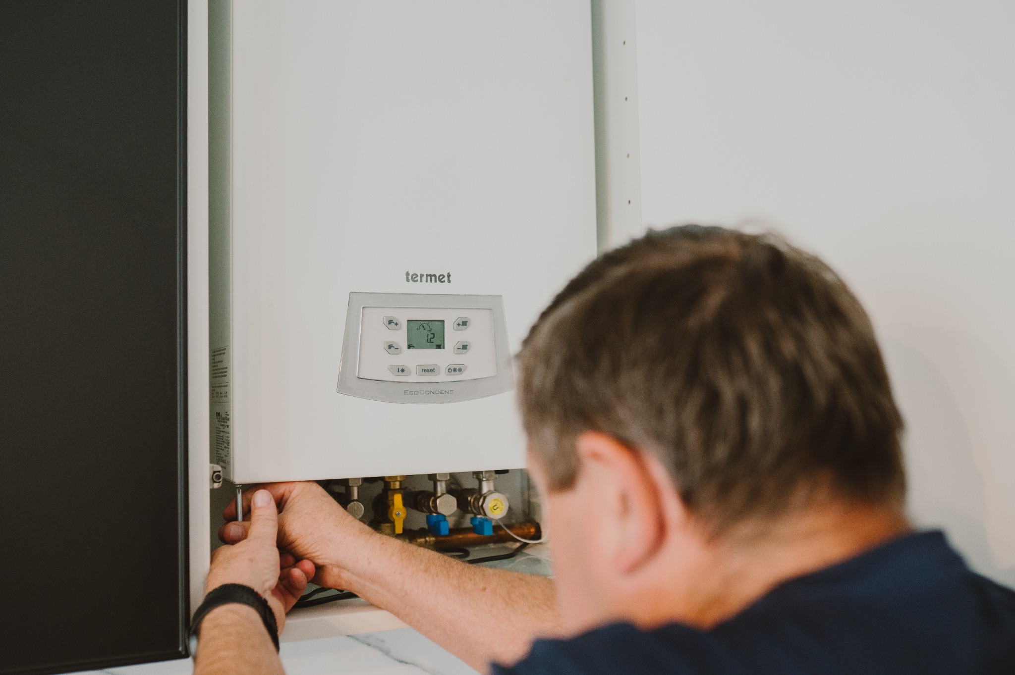 Mężczyzna serwisuje wiszący piec gazowy Termet. Widoczne zawory, wyświetlacz i dłonie montera z narzędziem.