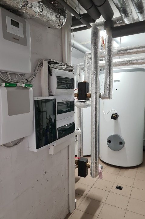 Białe zbiorniki, rury w srebrnej izolacji i skrzynki elektryczne w kotłowni. Widoczne przewody i instalacje.