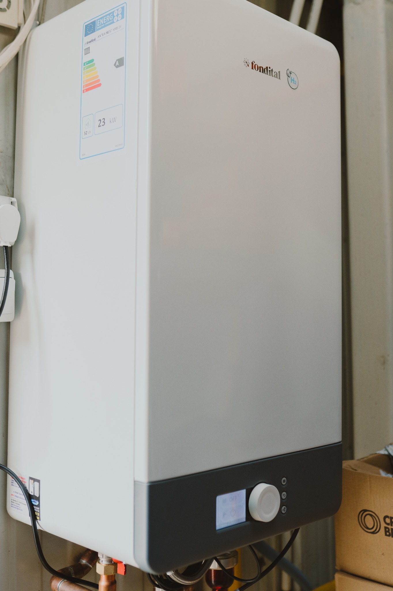 Biały, wiszący piec gazowy marki Fondital z etykietą energetyczną i widocznymi rurami doprowadzającymi, zamontowany na jasnej ścianie. Widoczny panel sterowania.