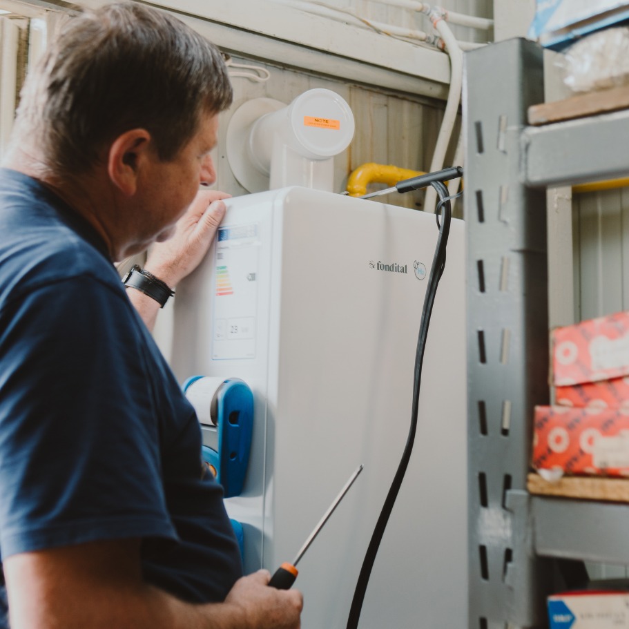 Instalator serwisuje piec gazowy marki Fondital, używając śrubokręta. Widoczna etykieta energetyczna i żółta rura gazowa. Ujęcie z boku, w tle regał magazynowy.